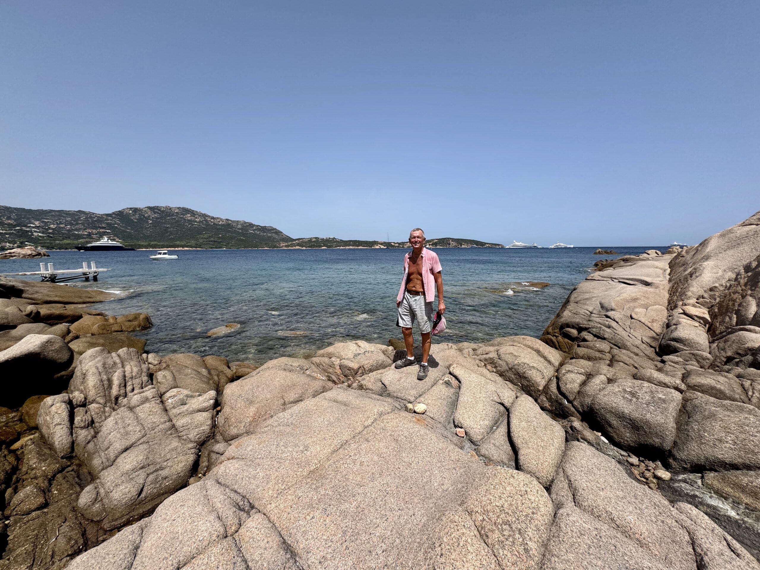 Man in a pink shirt standing on large rocky shore by calm blue water, with boats in the distance and hills on the horizon.