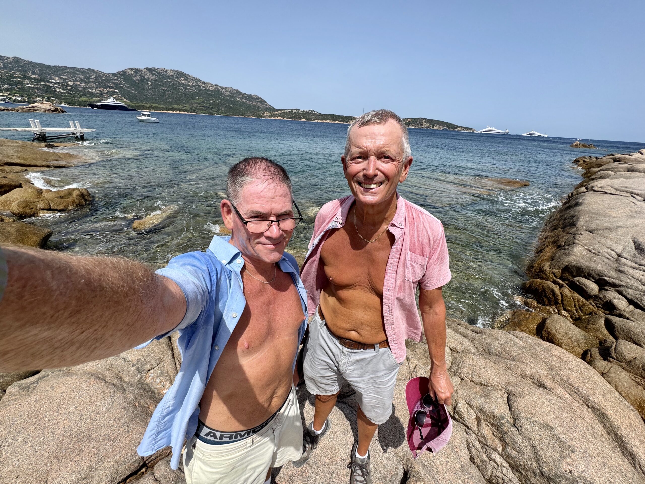 Two men posing for a selfie on a rocky seaside with clear blue water and hills in the background; several boats anchored offshore on a sunny day.