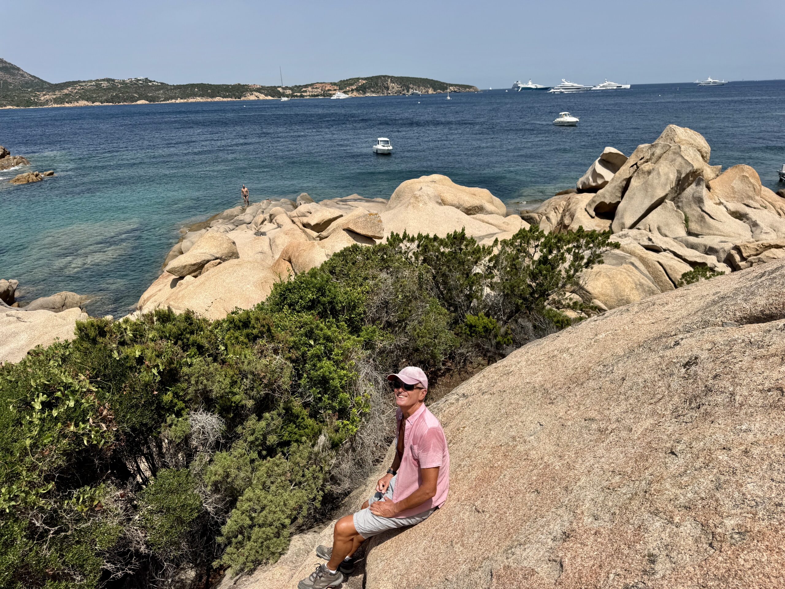 Person in a pink shirt and shorts climbs rocky coastline beside a blue sea with several boats in the distance.