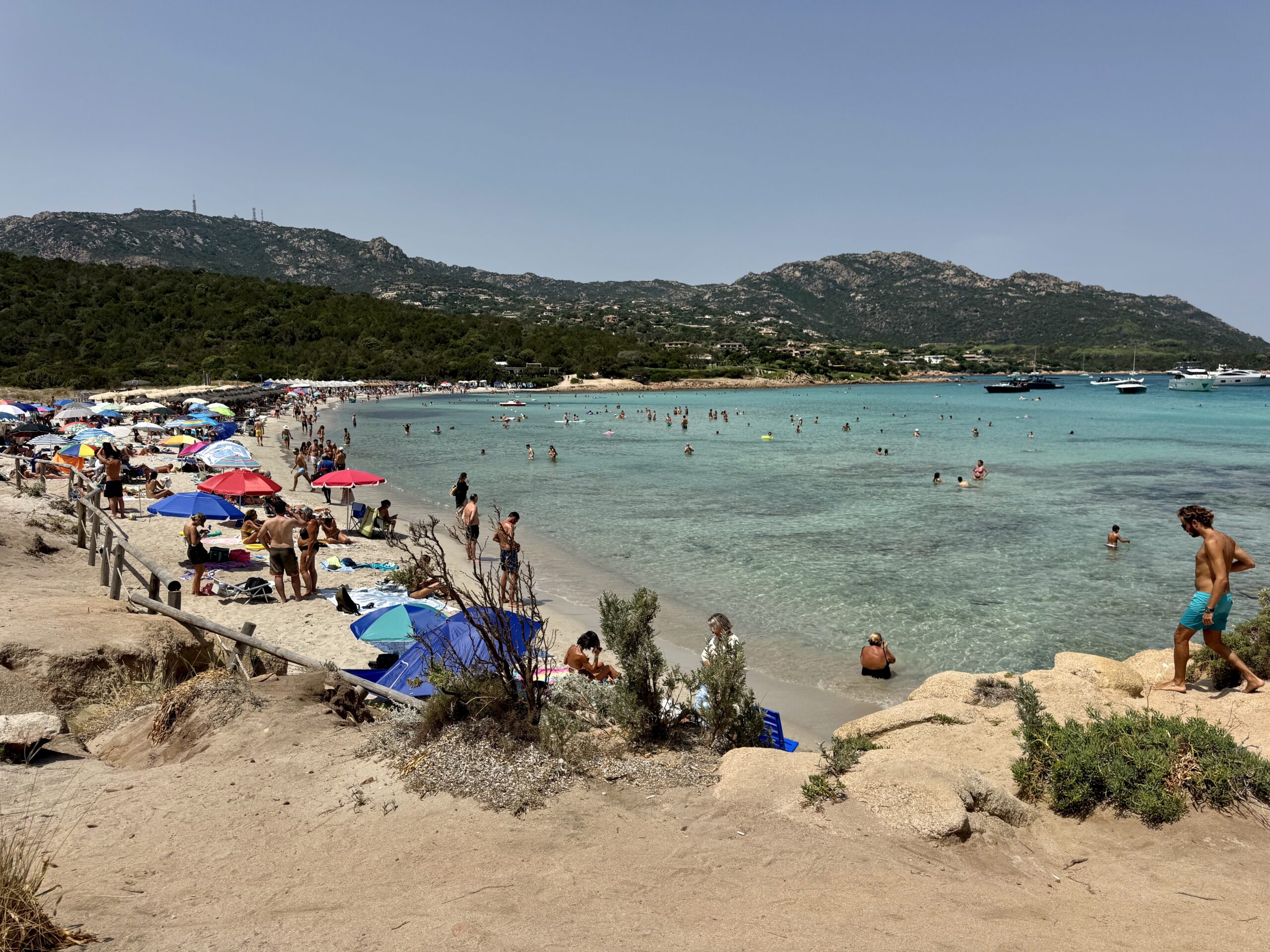 Crowded sandy beach along a turquoise bay with colorful umbrellas, sunbathers, and clear water, backed by green hills and mountains.