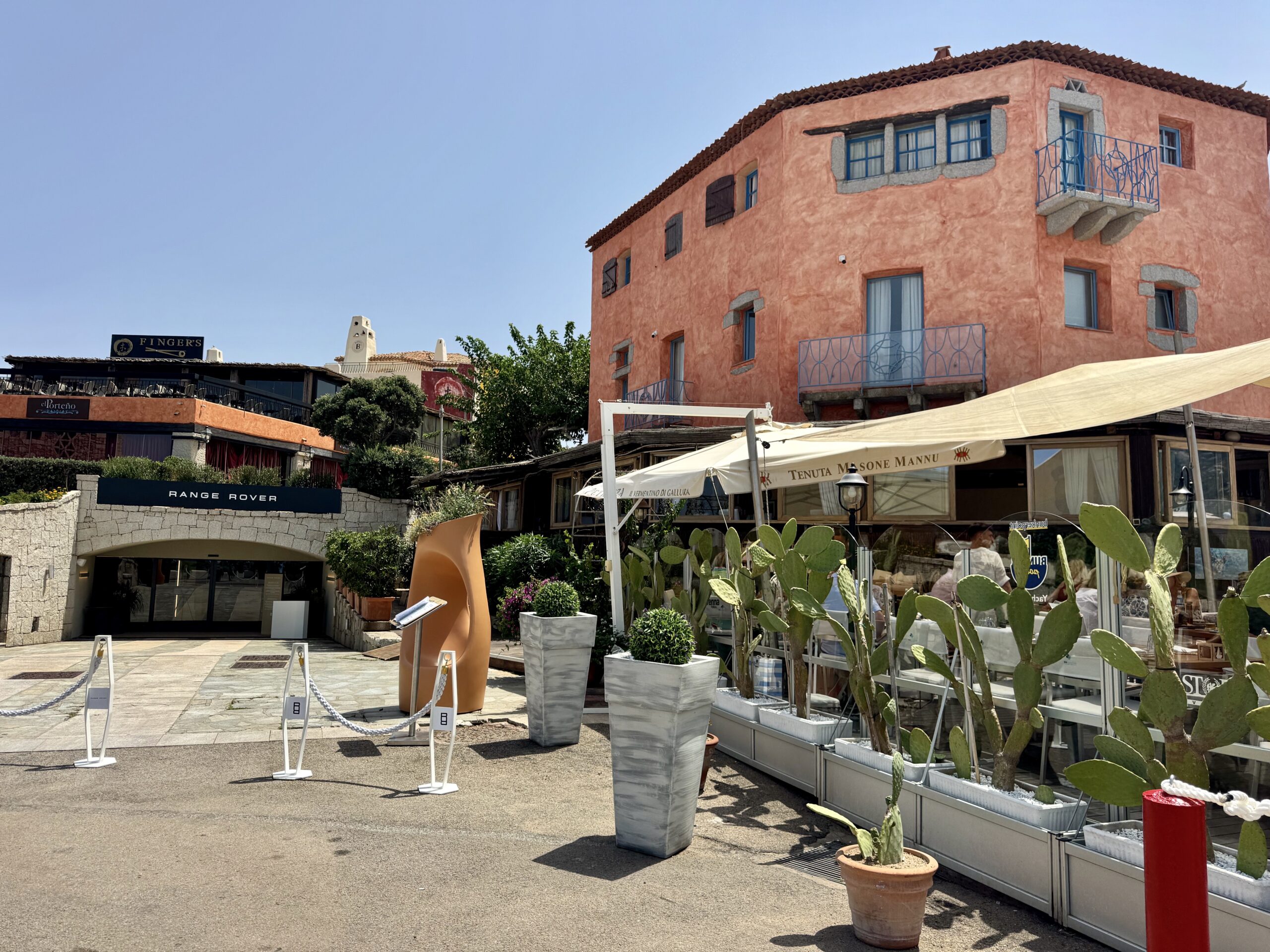 Outdoor plaza with a peach stucco building, blue balcony rails, and large potted cacti in the foreground.