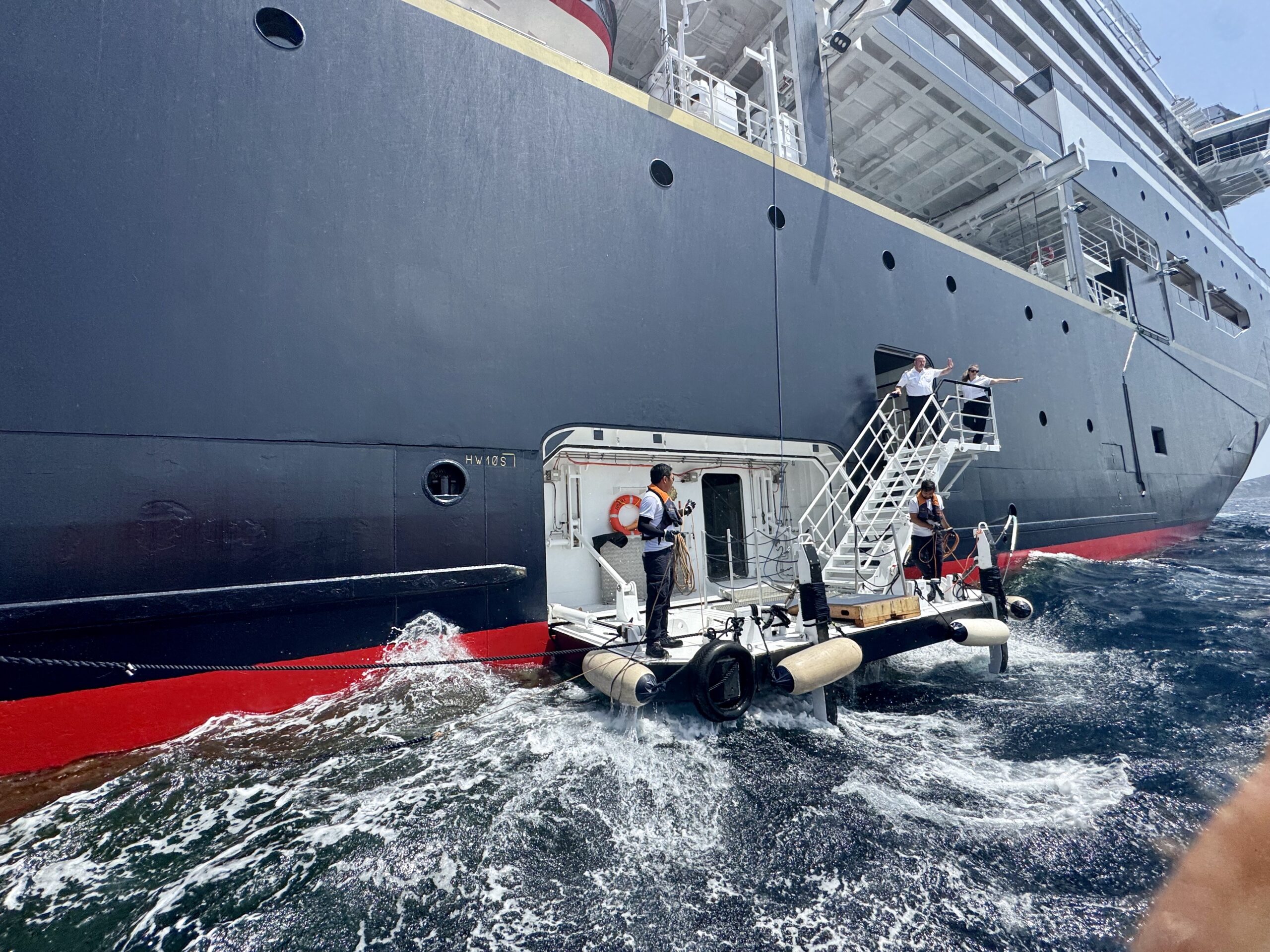 Crew in life vests standing on a small tender boat alongside a large gray ship at sea during a docking maneuver.