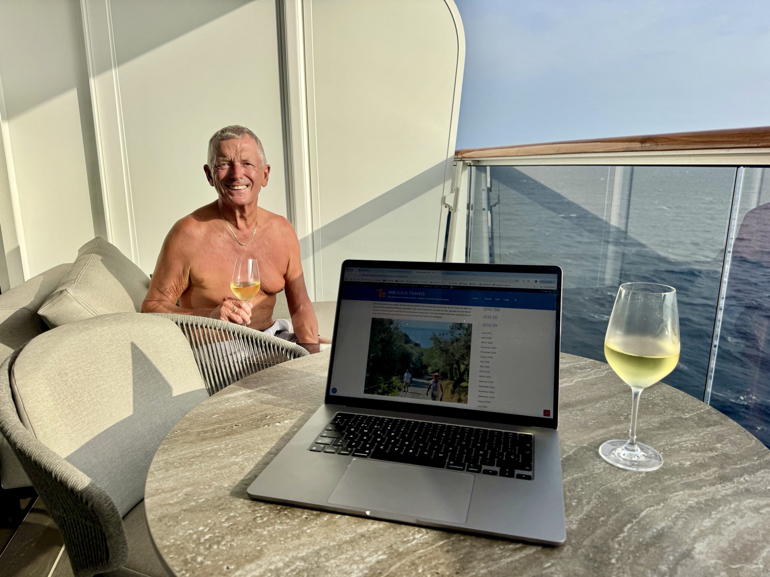 Smiling shirtless man sitting on a boat deck, holding a glass of white wine with a laptop nearby and another wine glass on the table against a sea backdrop.
