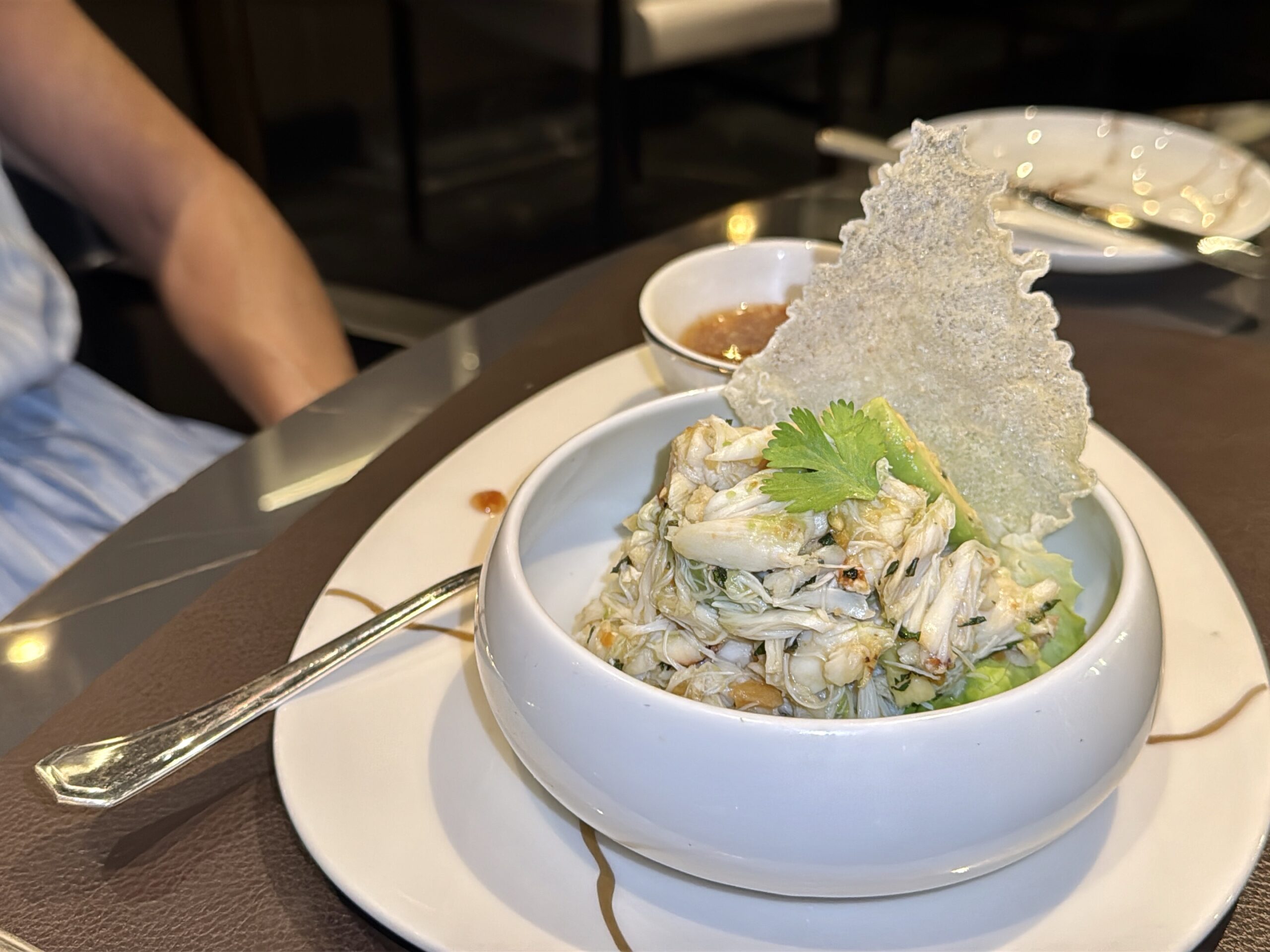 Bowl of shredded cabbage salad with herbs, topped by a crispy lacy cracker, beside a small bowl of sauce on a white plate and table.