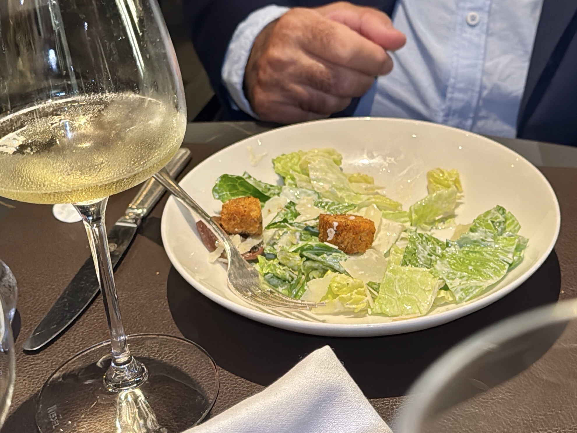 Caesar salad with romaine and croutons in a white bowl, along with a glass of white wine on the table and a hand in the background.