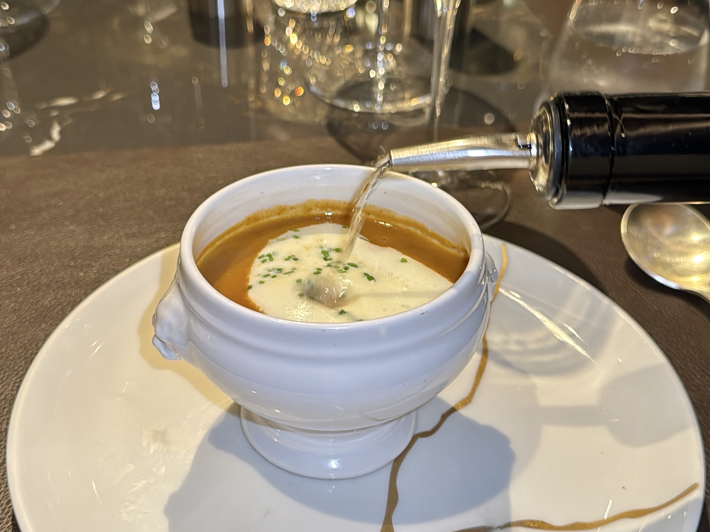 Cup of orange creamy soup with a cream swirl and green herbs in a white cup on a saucer; liquid being poured into the cup from a bottle nearby.