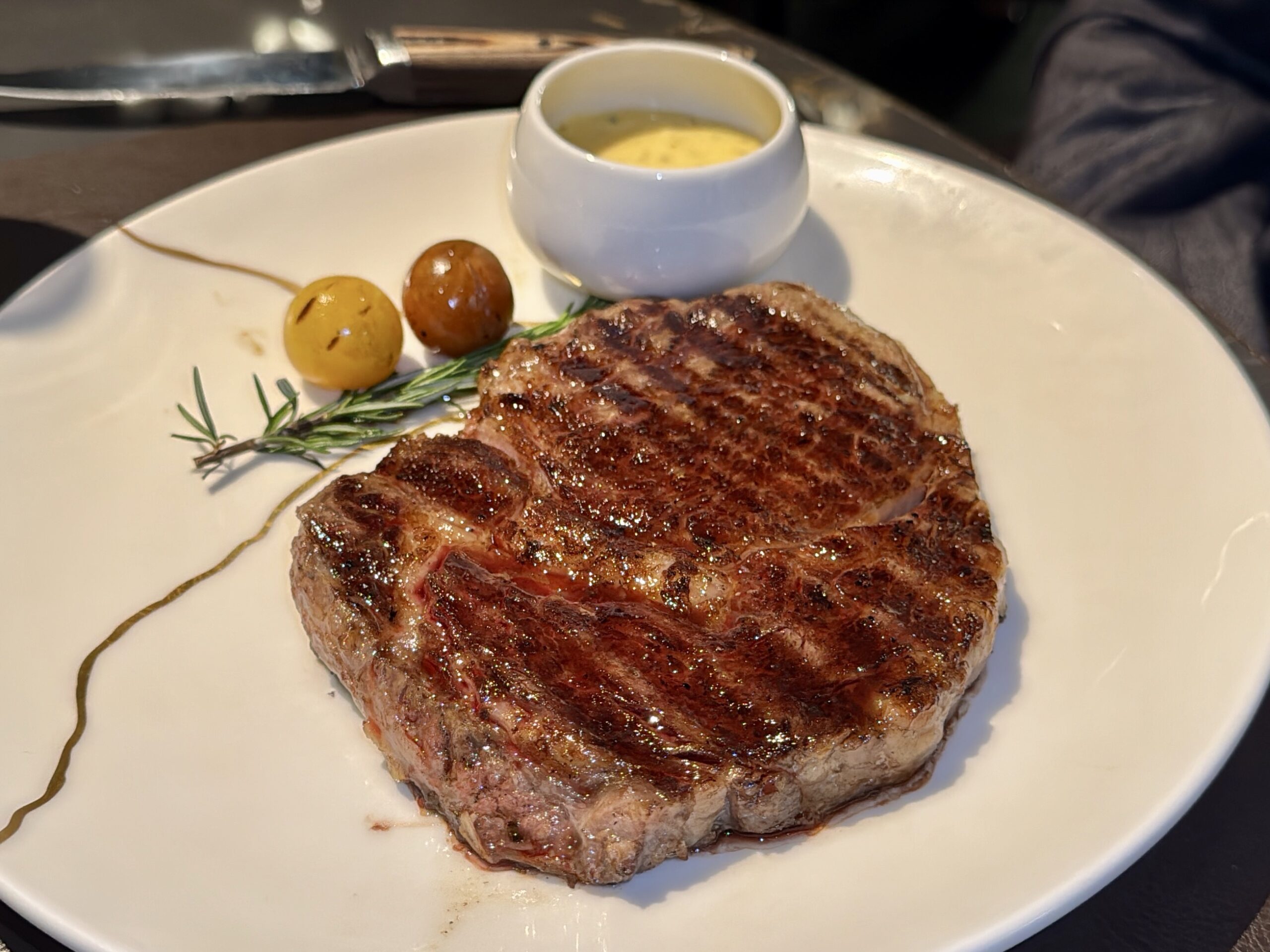 Juicy grilled steak on a white plate with rosemary, two olives, and a small ramekin of sauce nearby.
