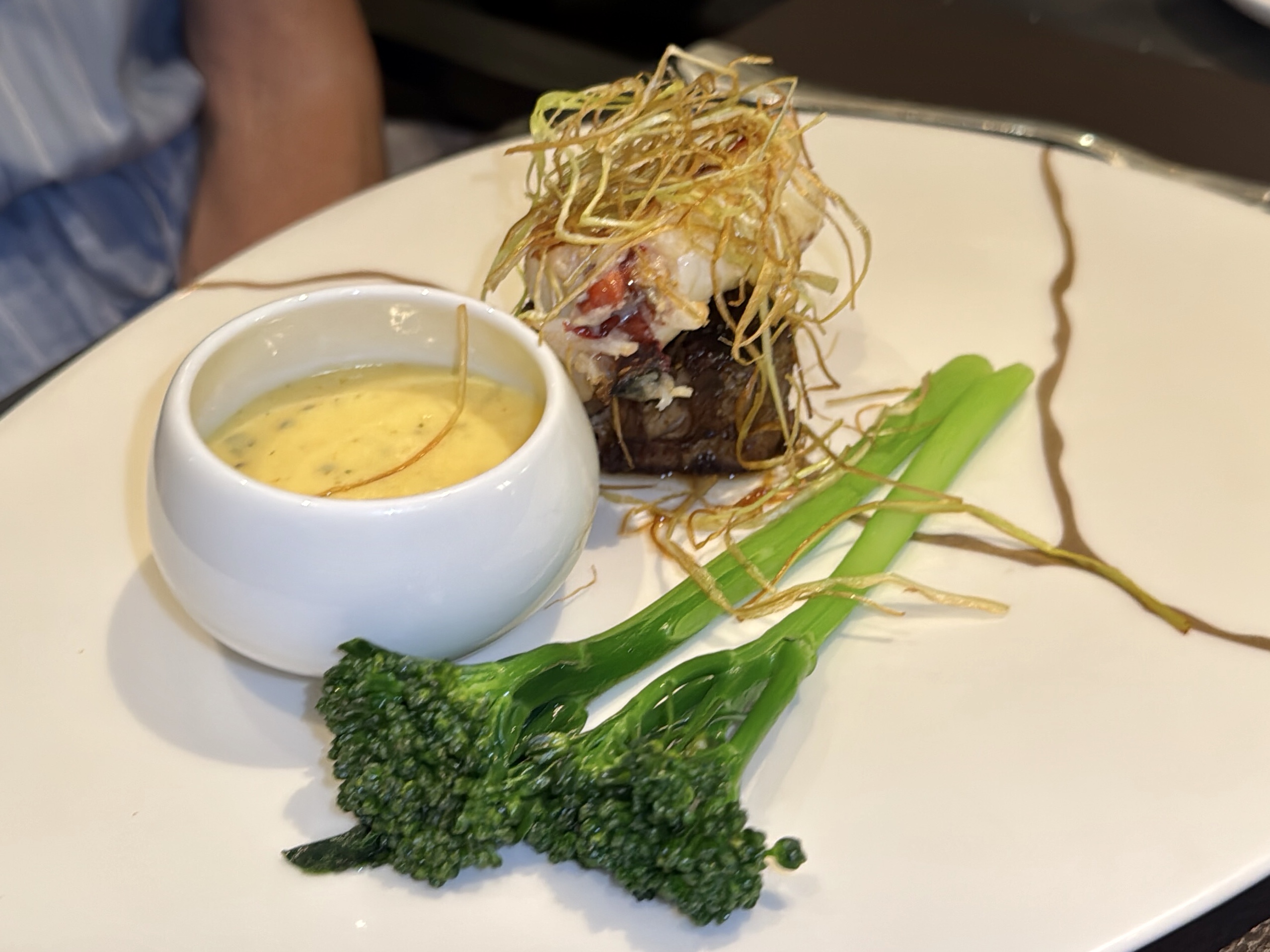 Plate with a meat entrée topped by a nest of crispy fried onions, accompanied by a small bowl of creamy dipping sauce and green vegetables on the side.