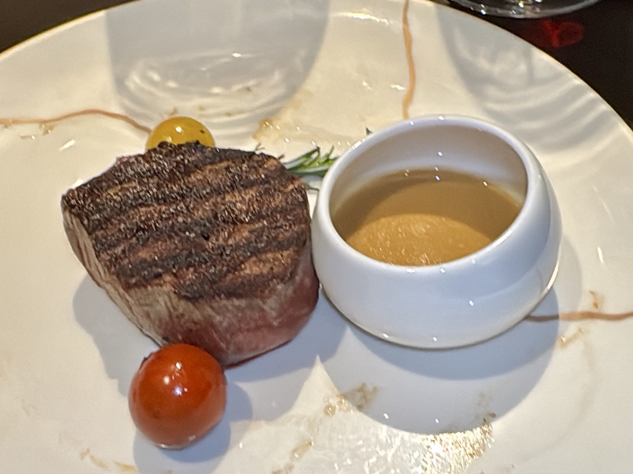 Grilled steak on a white plate with a small cup of sauce and two glossy cherry tomatoes; plate has sauce splatters and herbs in background.