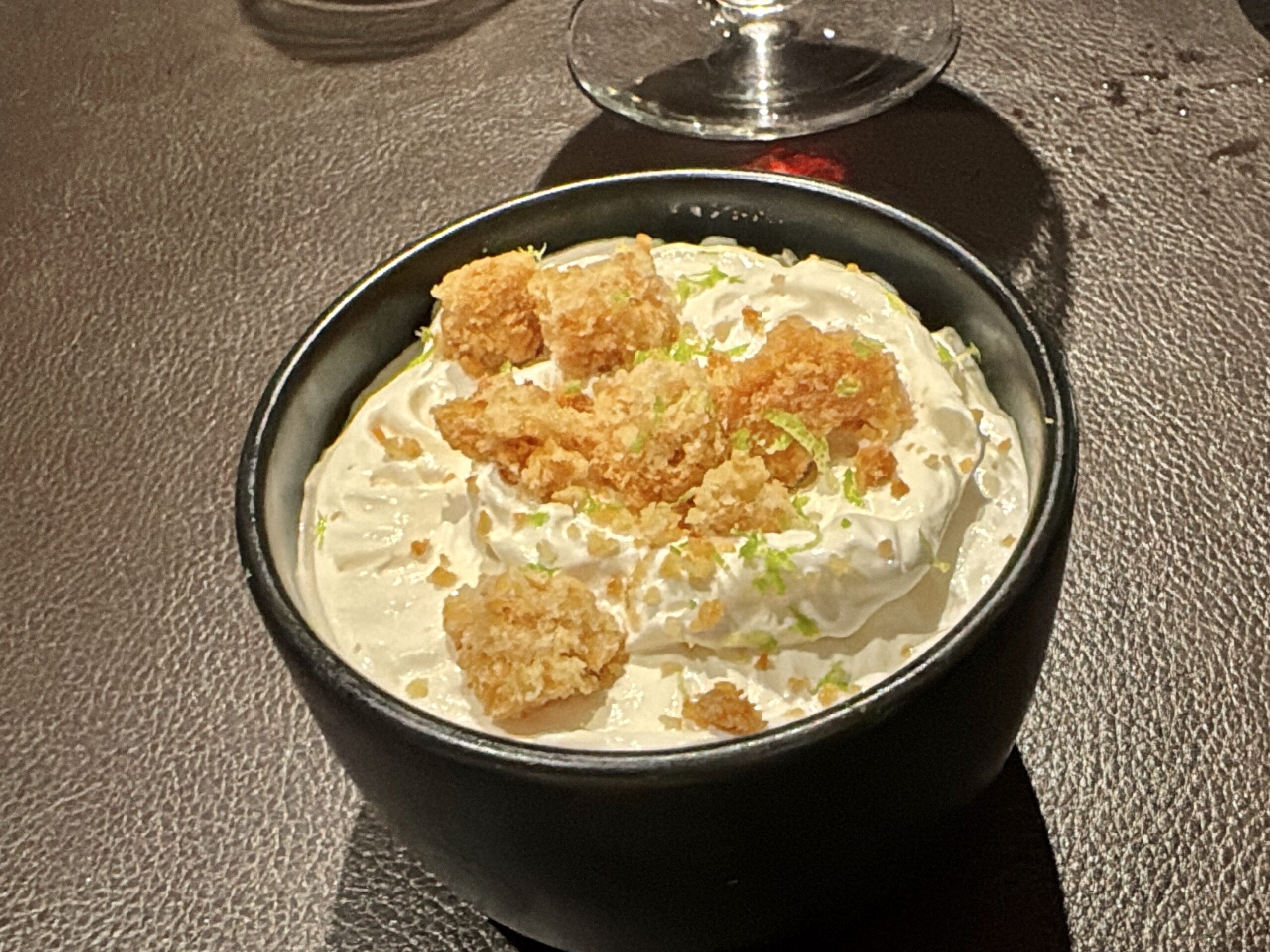 Small black bowl with a creamy dessert topped by crispy fried crumbles and green herbs on a dark textured table surface.