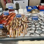 Assorted whole fish laid on crushed ice at a market counter with blue price cards, including red-orange reef fish and silver-grey fish on the right.