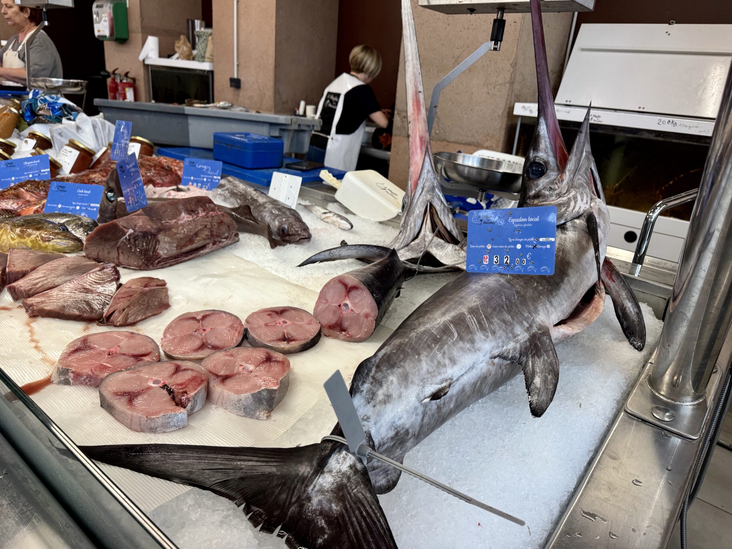 Seafood market stall displaying large tuna and tuna steaks on crushed ice with blue price signs; a worker in apron is visible in the background.