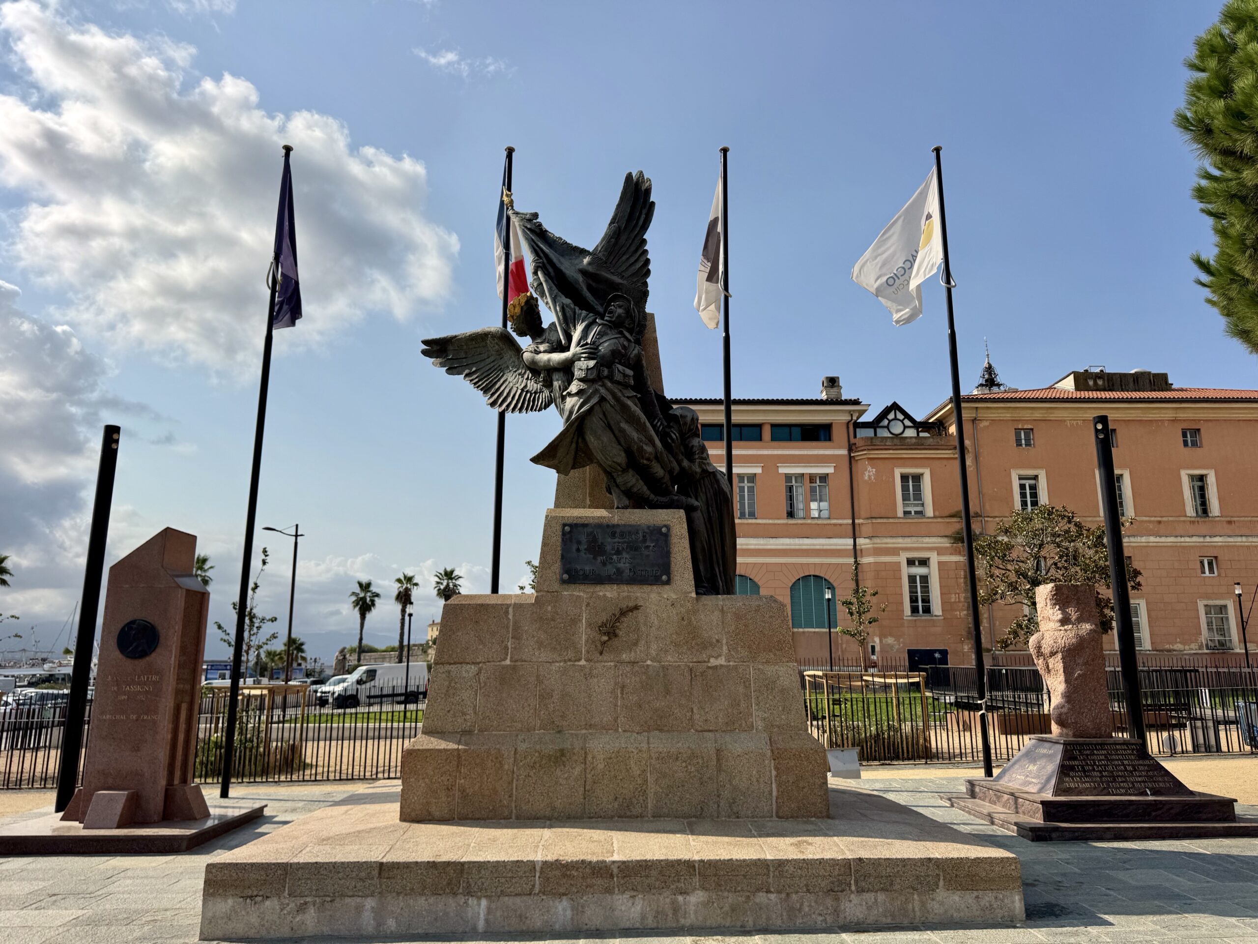 Bronze angel statue with outstretched wings on a stone pedestal, flanked by flagpoles and a peach-colored building in the background under a blue sky