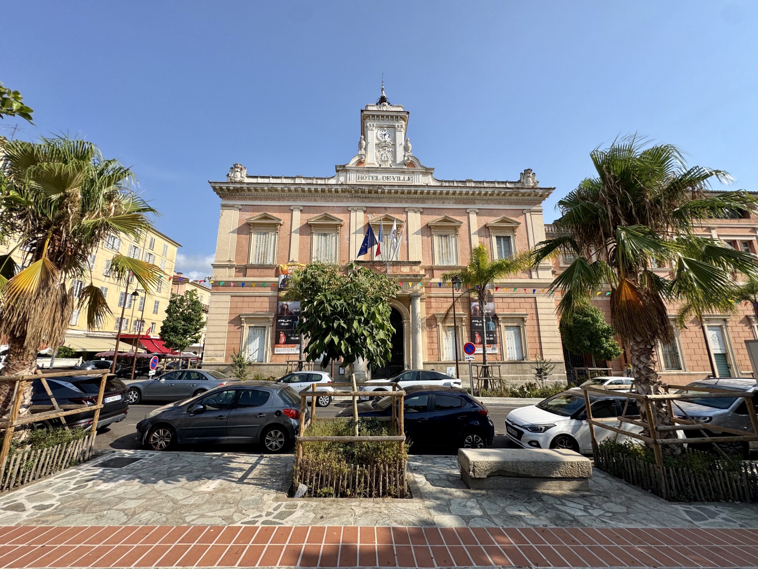 Facade of Hôtel de Ville with a clock tower rising above a pinkish-beige town hall, palm trees framing the plaza on a sunny day