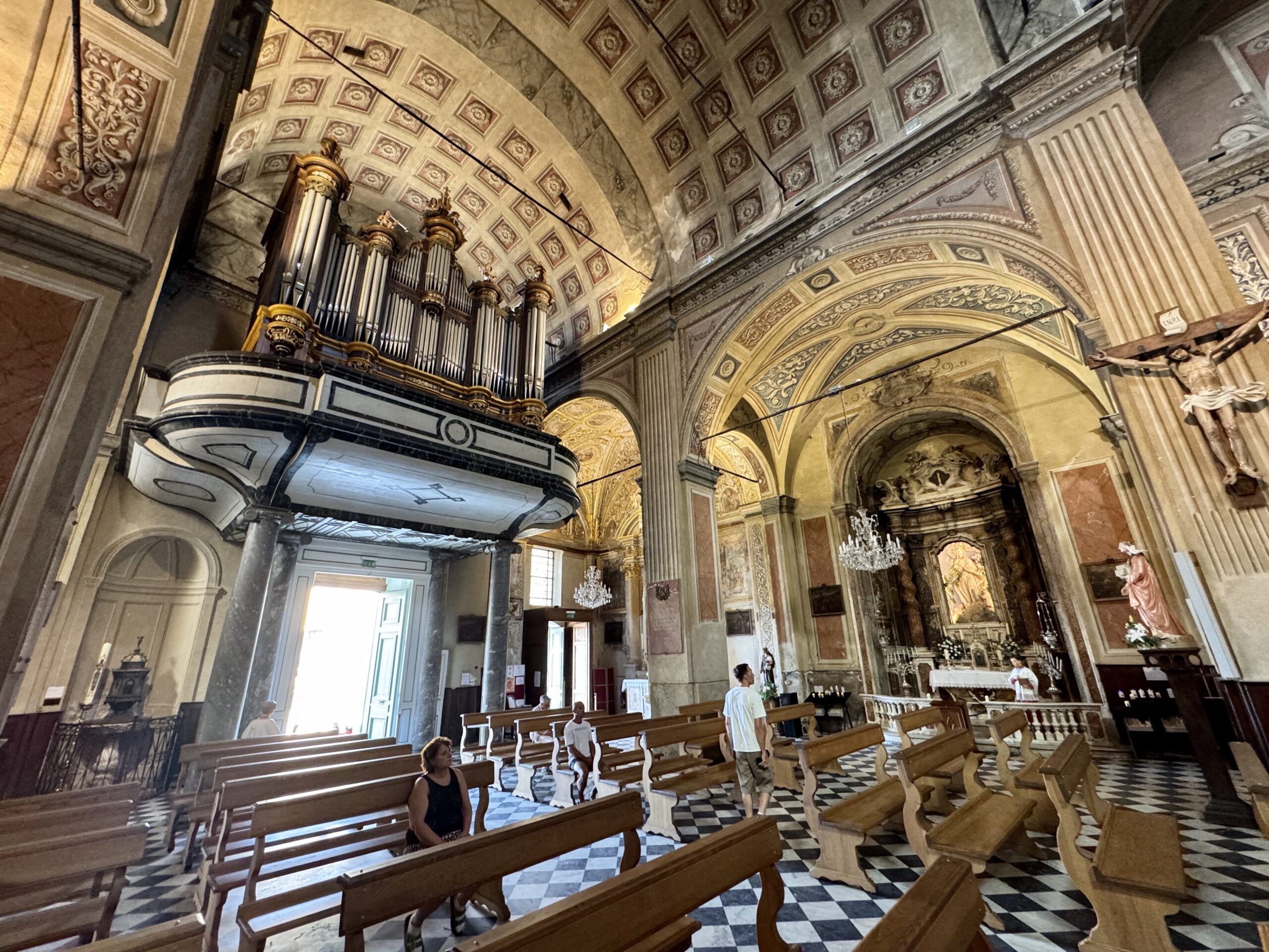 Interior of ornate church with an elevated pipe organ and wooden pews, checkered floor, and carved arches.
