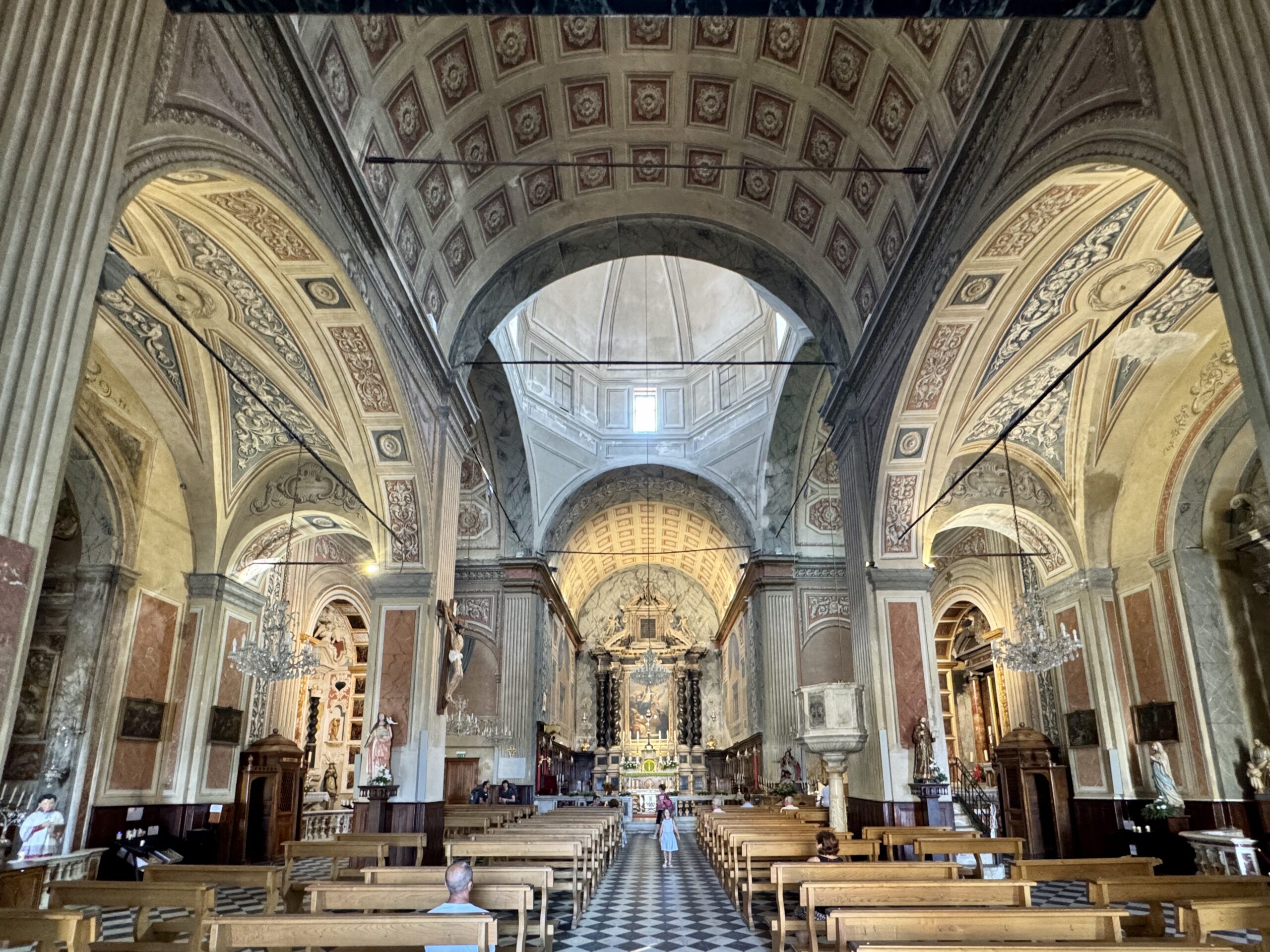 Interior of a grand cathedral with ornate vaulted ceilings, arches, and rows of wooden pews leading to an ornate altar at the far end.