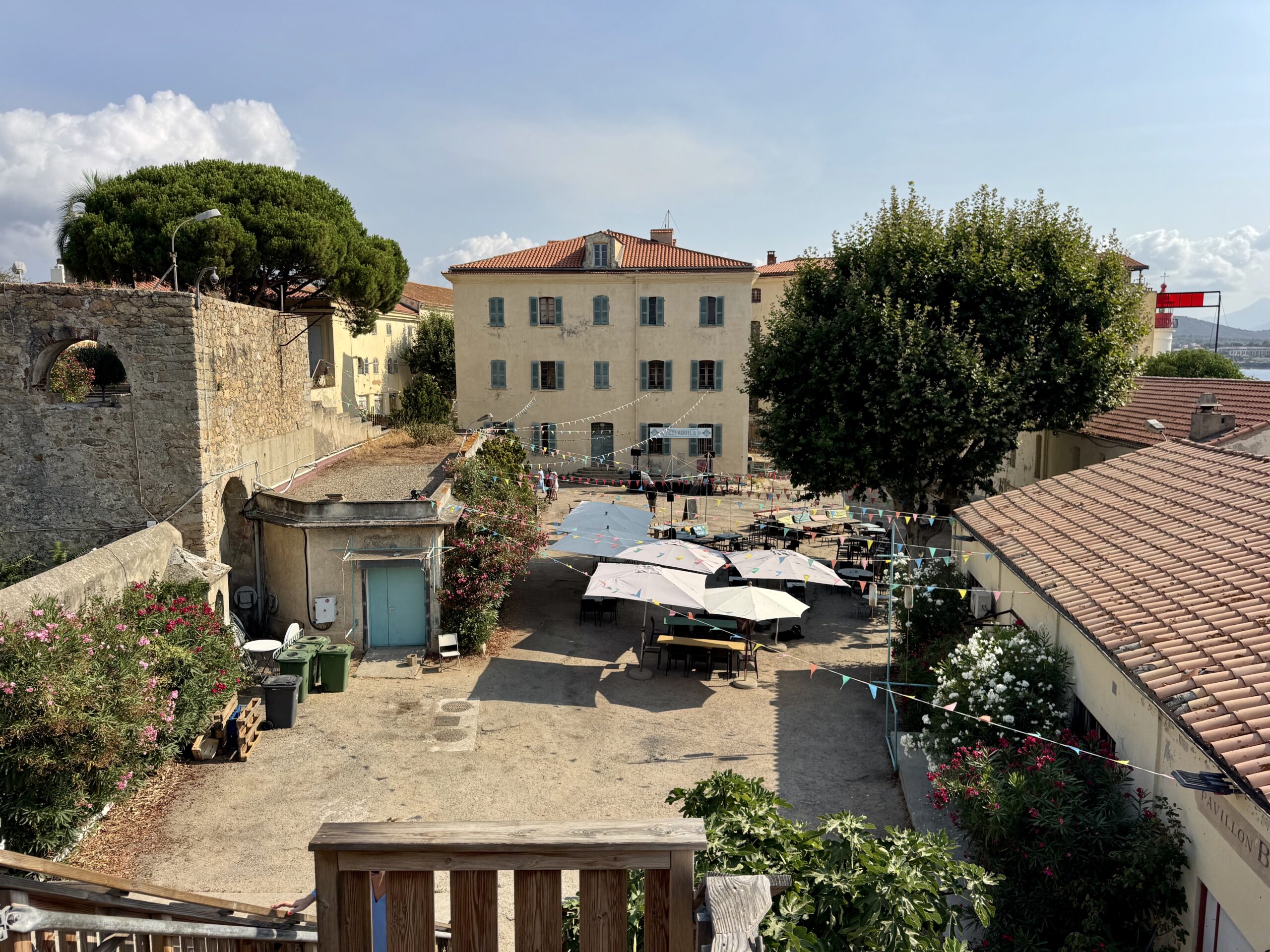 Sunlit village courtyard with cream buildings, blue shutters, and outdoor tables under white umbrellas.","Cobbled alleyway between stone walls and pastel houses, string bunting and cafe seating.","Town square with bunting, umbrellas, and tables, bordered by red-tiled roofs and greenery.","Rooftops, trees, and a sunny plaza with chairs and plants in a coastal village."