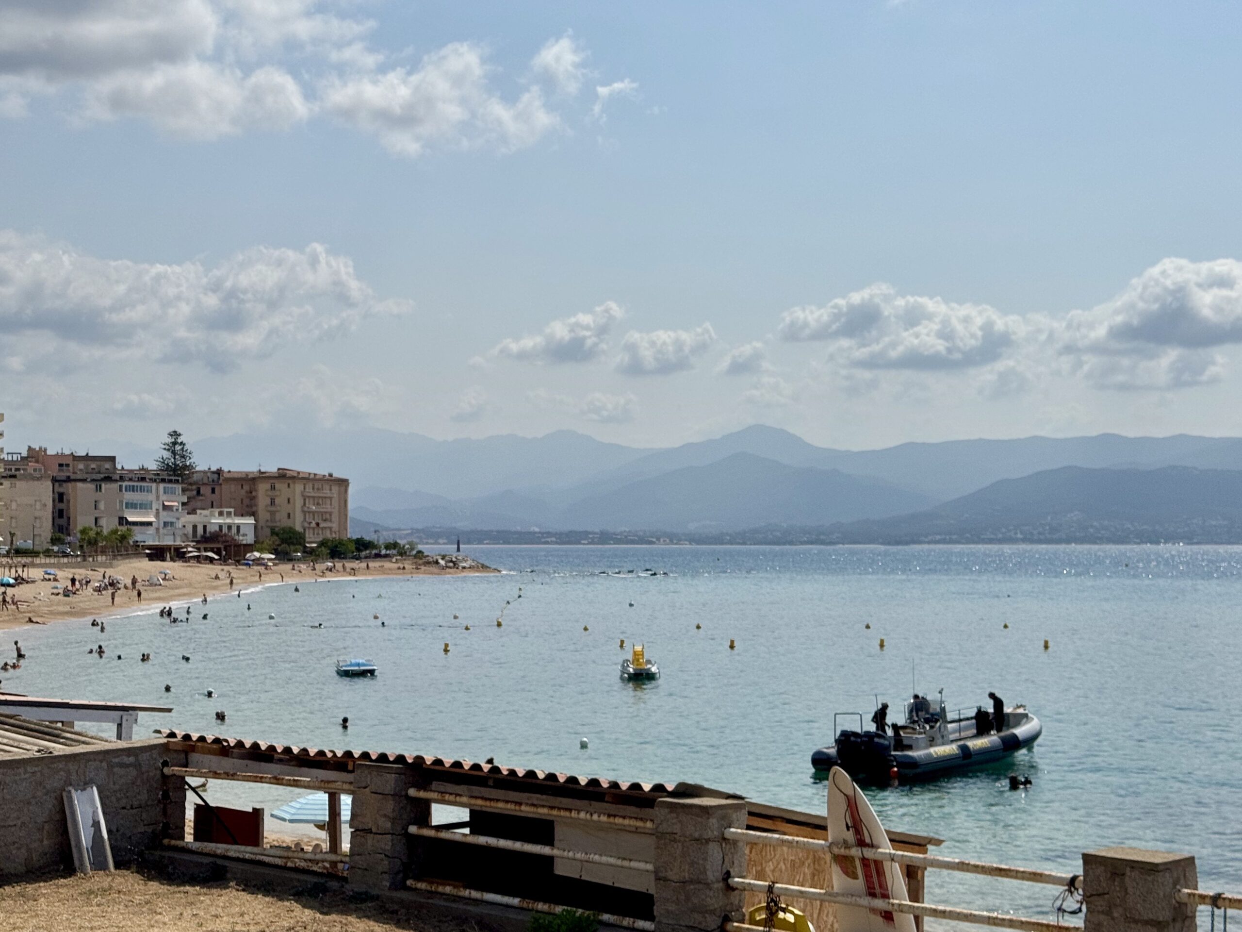 Coastal beach town with buildings along the shore, people in the water, and distant blue mountains under a partly cloudy sky.
