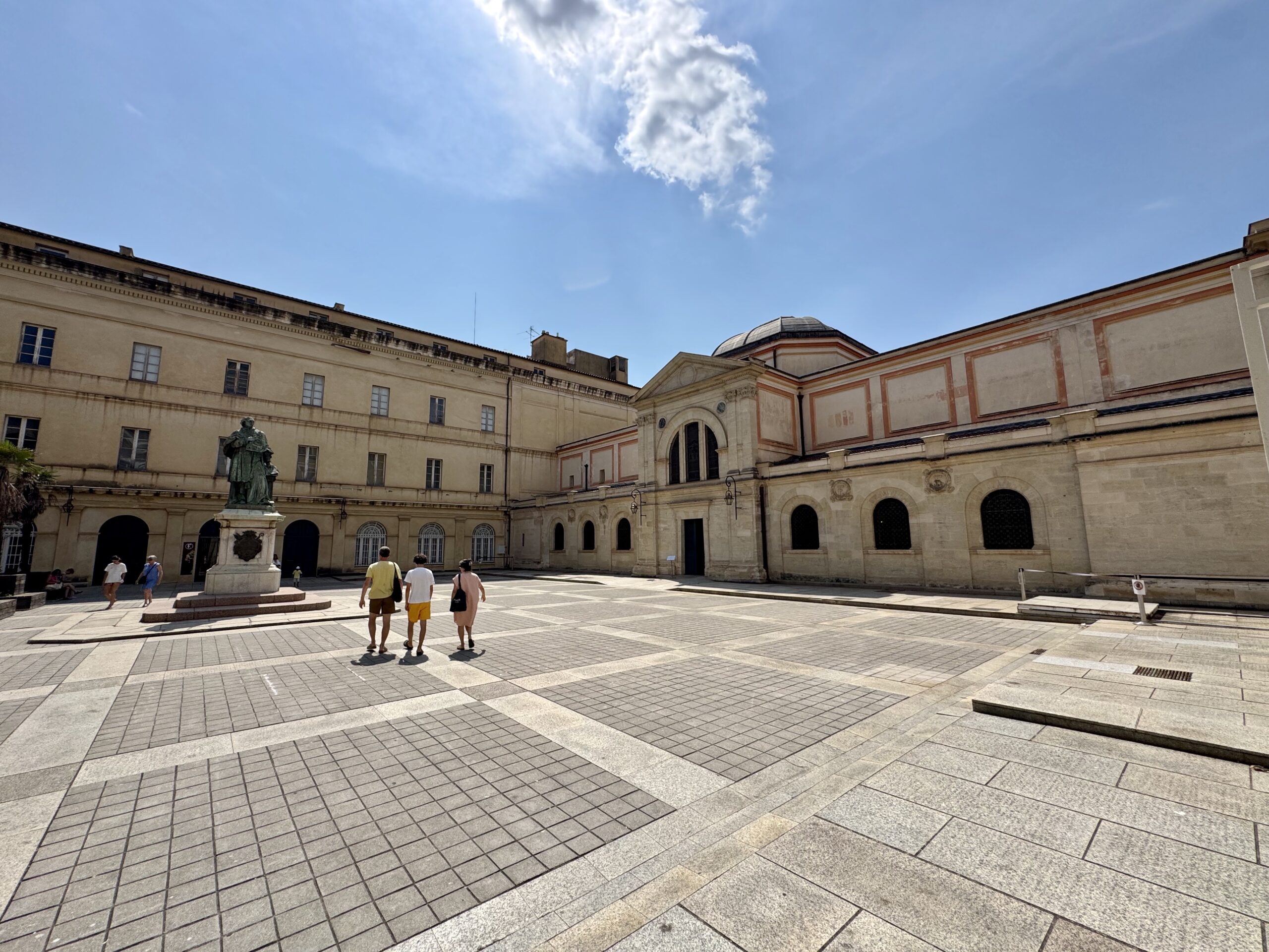 Statue in a tiled European plaza with beige historic buildings and a bright blue sky in the background, a few people walking nearby.