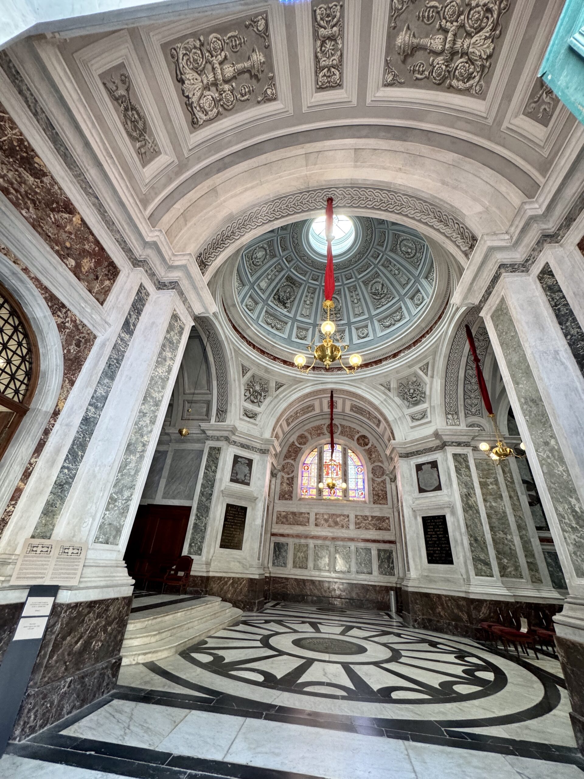 Interior of a grand marble church with ornate vaulted ceiling, circular dome, and gold chandelier visible above a black-and-white patterned floor.
