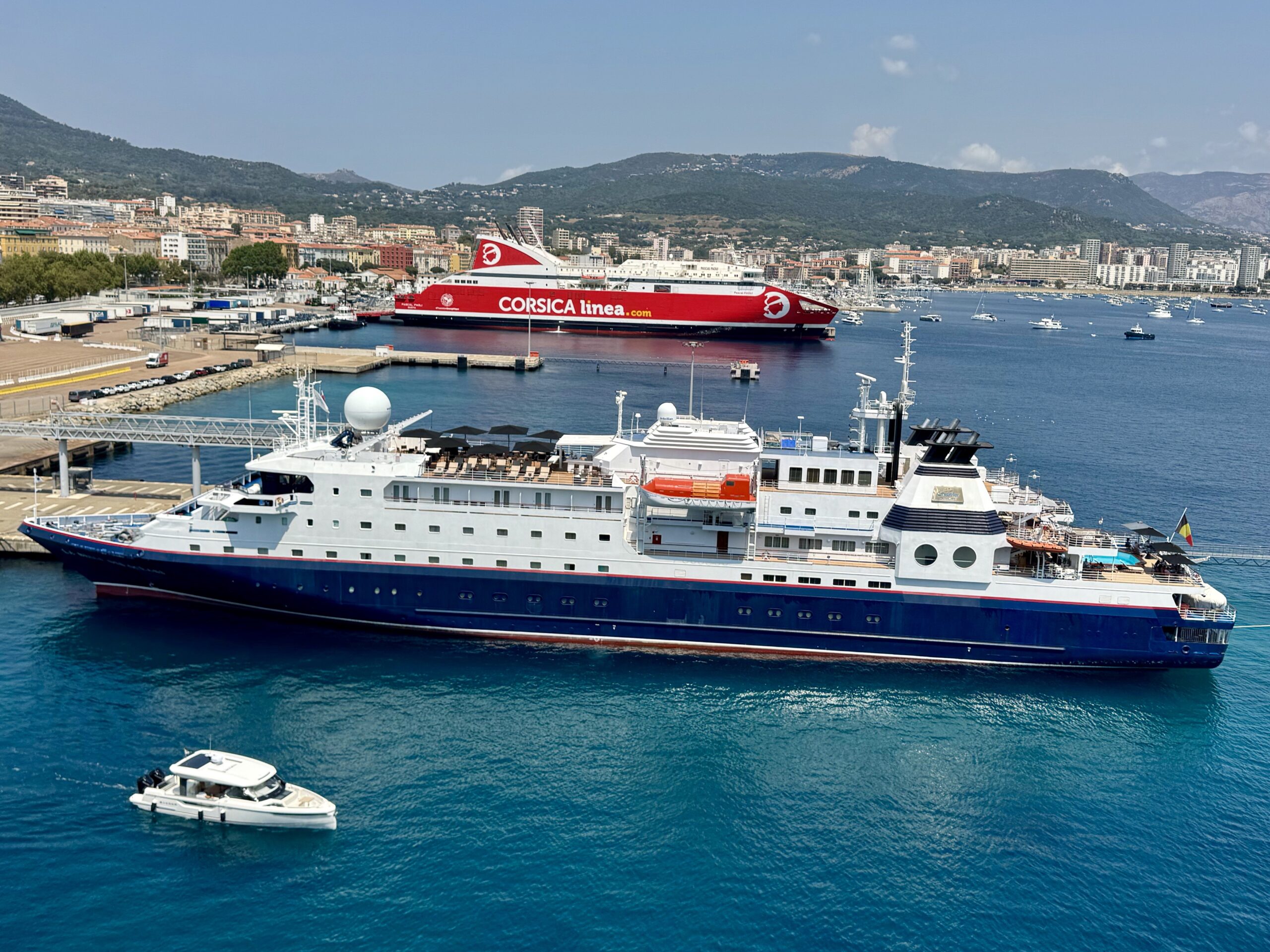 Red Corsica Linea passenger ferry docked in a harbor, with a coastal town and green hills in the background.