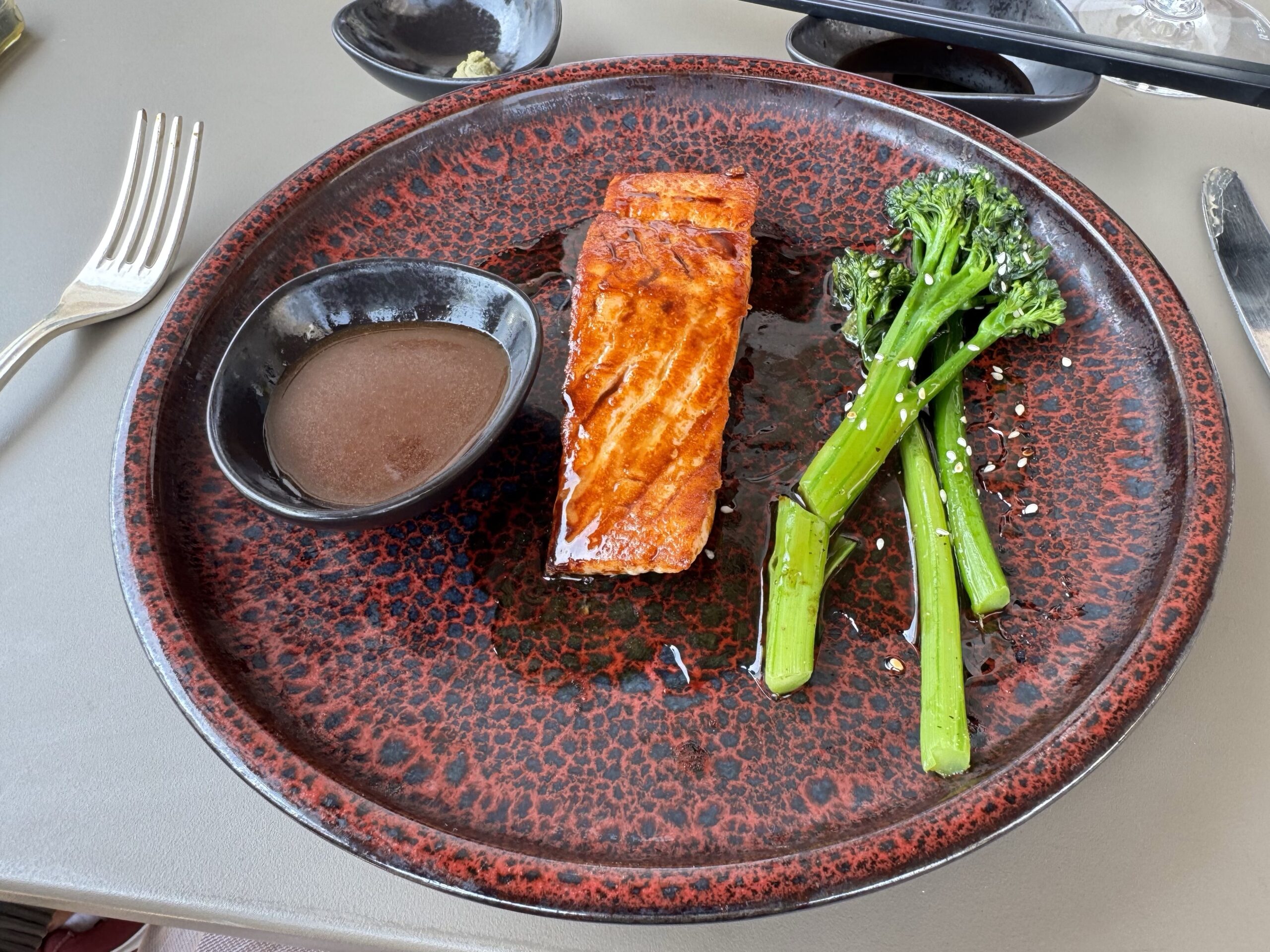 Grilled salmon fillet with broccoli on a red speckled plate and a small sauce cup. A fork and knife are nearby.