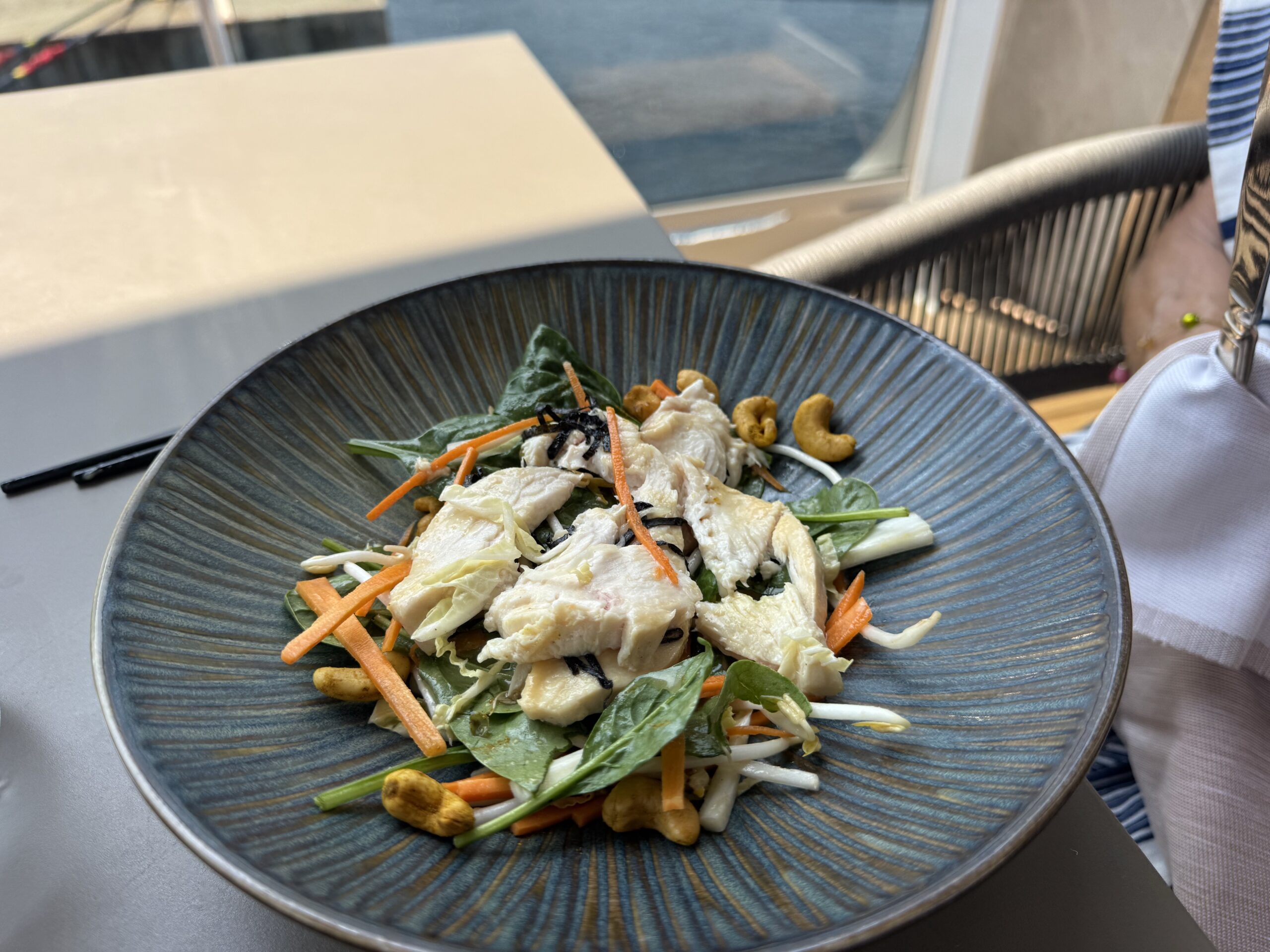 Blue ceramic bowl filled with chicken salad, shredded carrot, greens, and nuts on a light table outdoors.