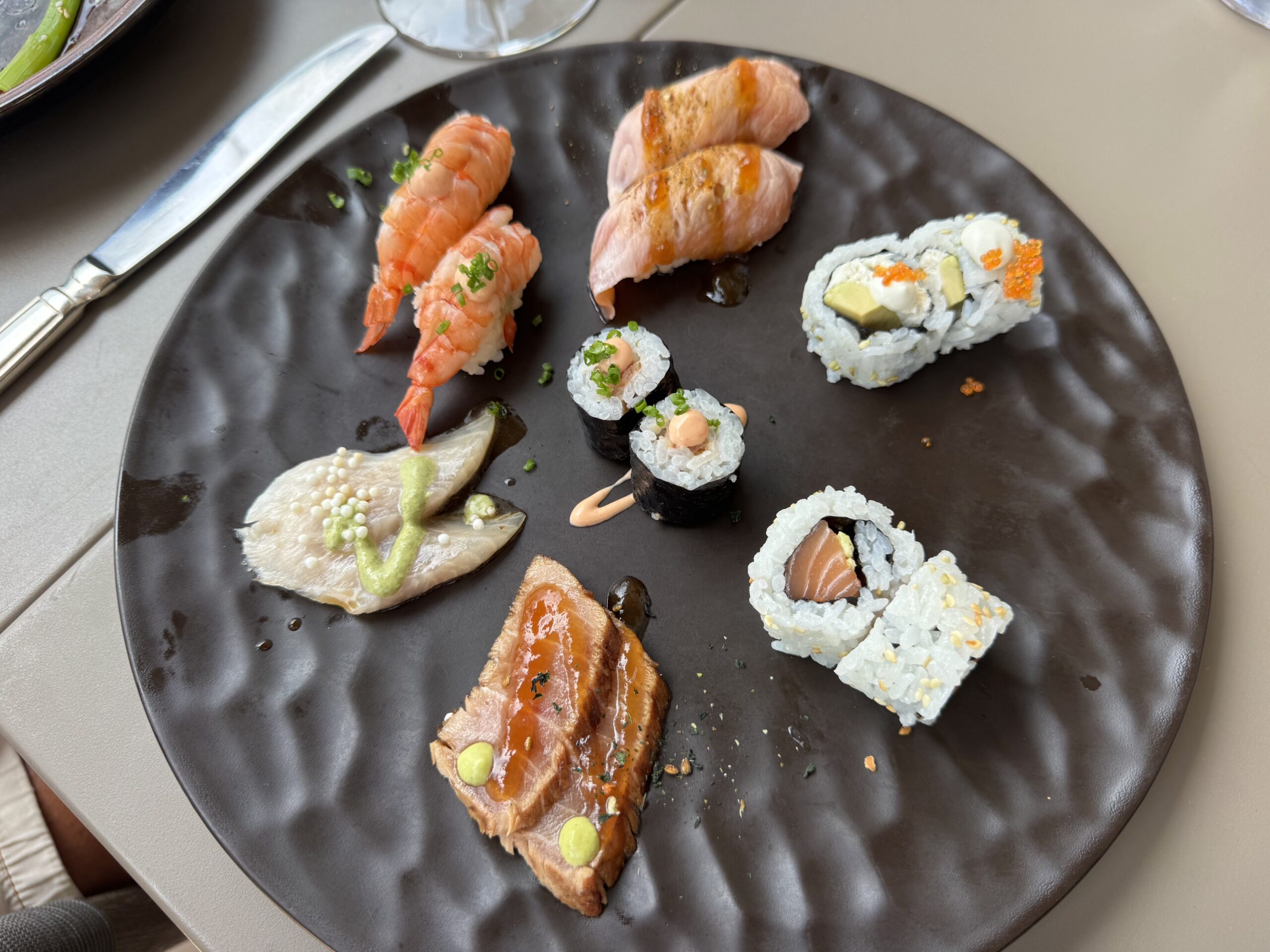 Plate of assorted sushi on a dark plate: shrimp nigiri, glazed salmon nigiri, and assorted tuna/rolls with avocado and orange roe; a few green onion garnishes add color.