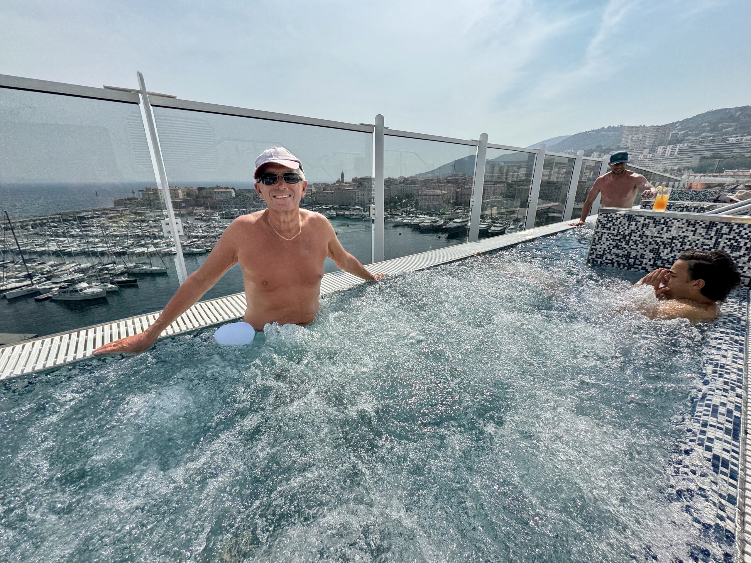 Two shirtless men enjoy a rooftop pool with glass railing, overlooking a marina and hillside cityscape.