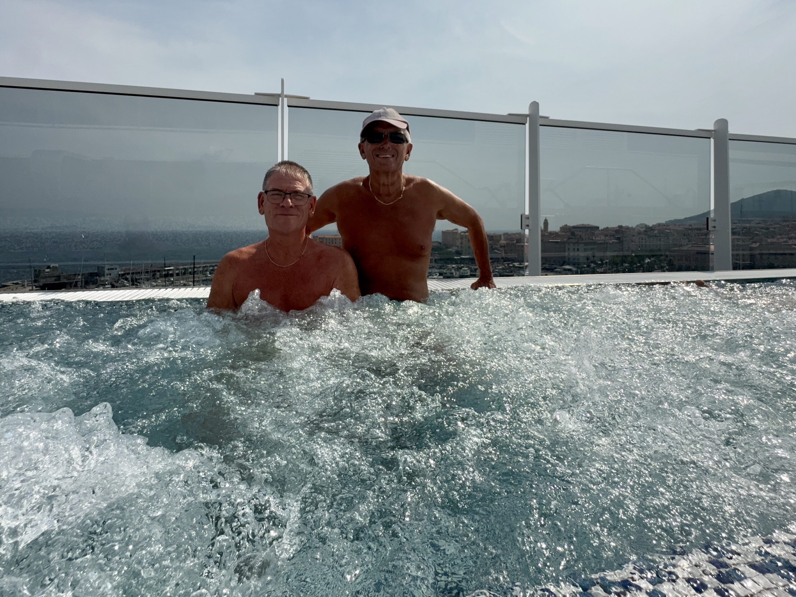 Two shirtless men posing in a rooftop pool, with a glass railing and a coastal cityscape in the background.