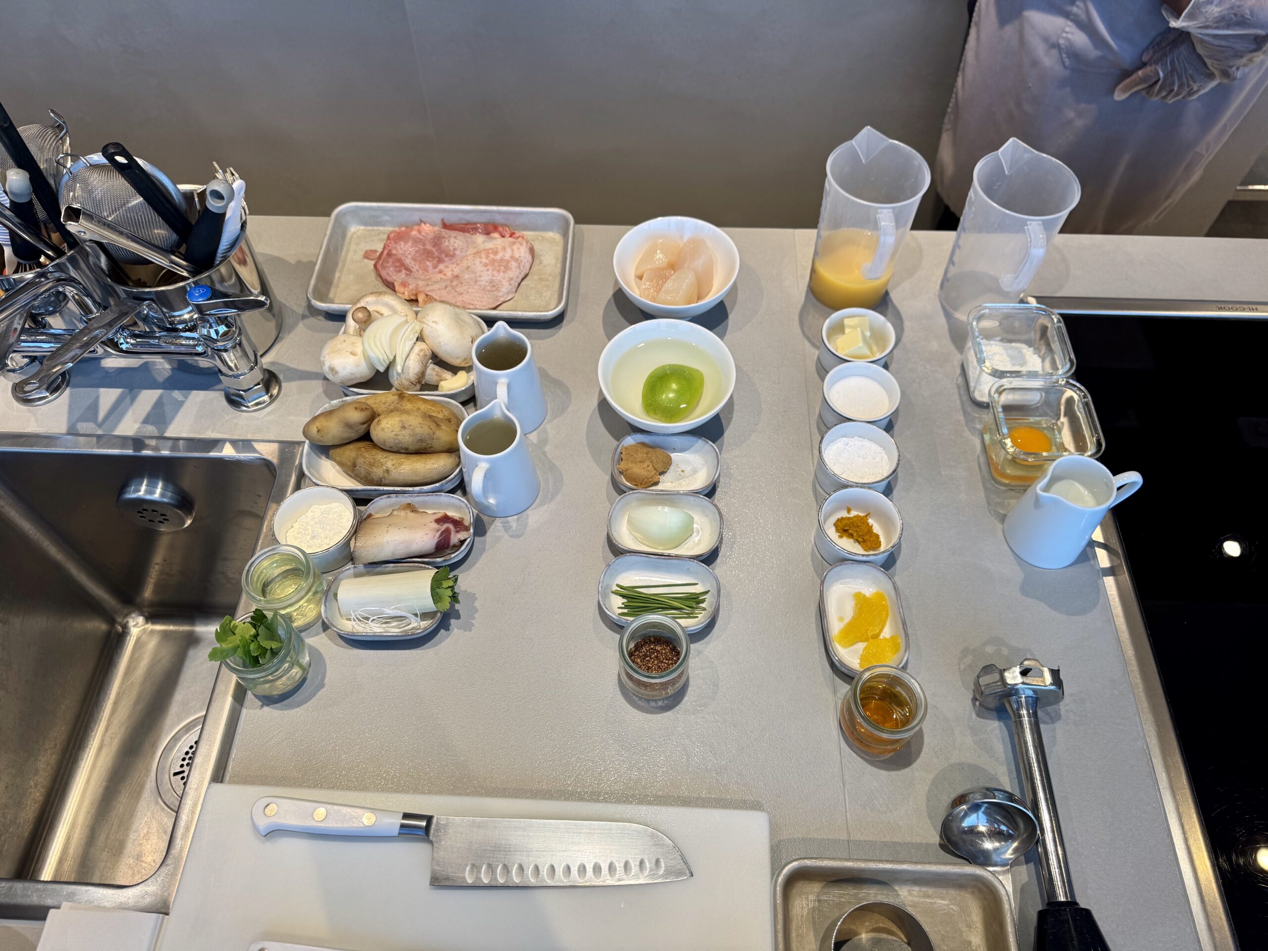 Professional kitchen prep station with raw meat on a tray, mushrooms, onions, lime, butter, oils, and bowls of spices and sauces laid out on a stainless counter