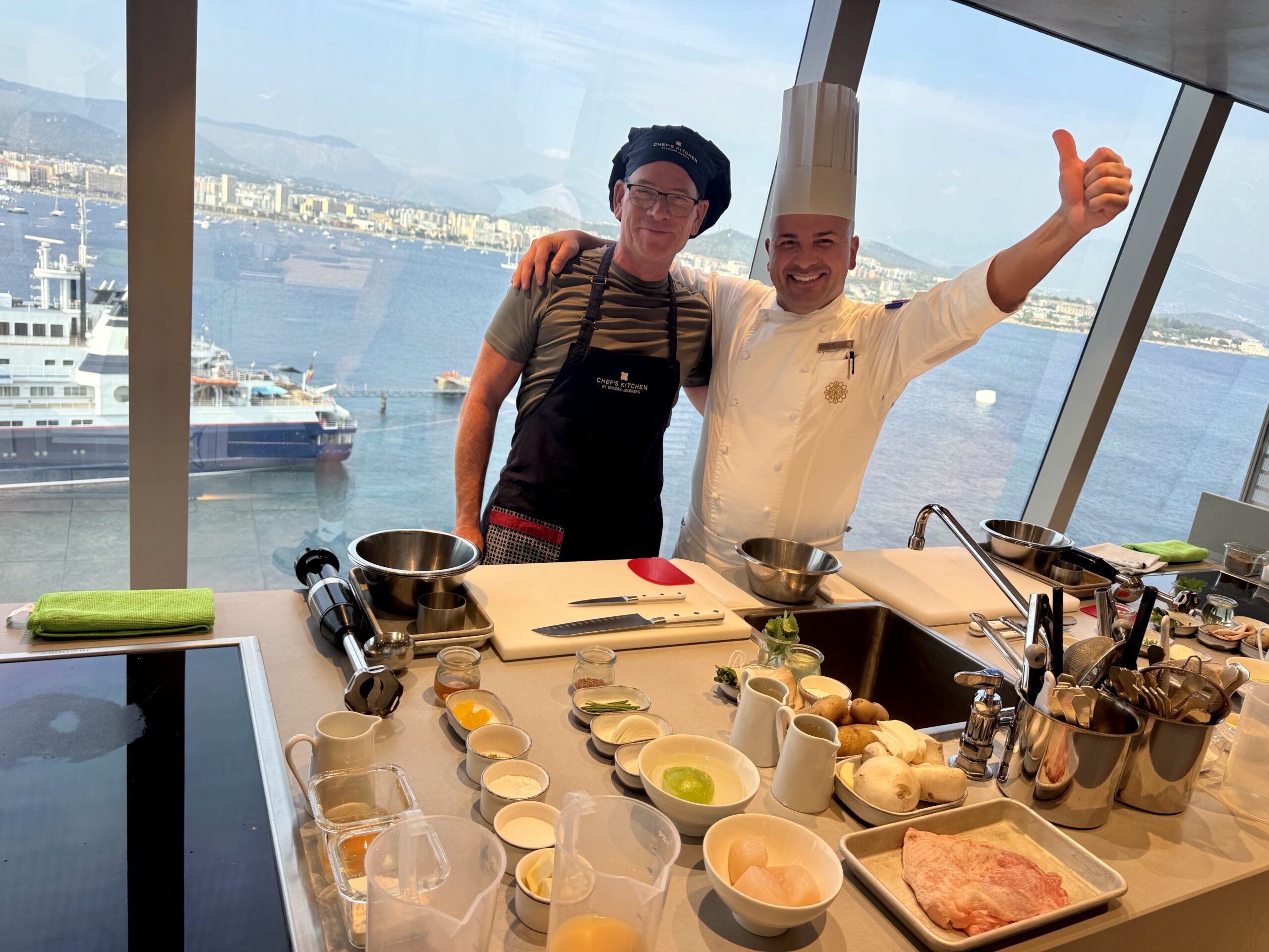 Two chefs pose at a bright kitchen counter with bowls of ingredients, one giving a thumbs-up and the harbor view behind them.