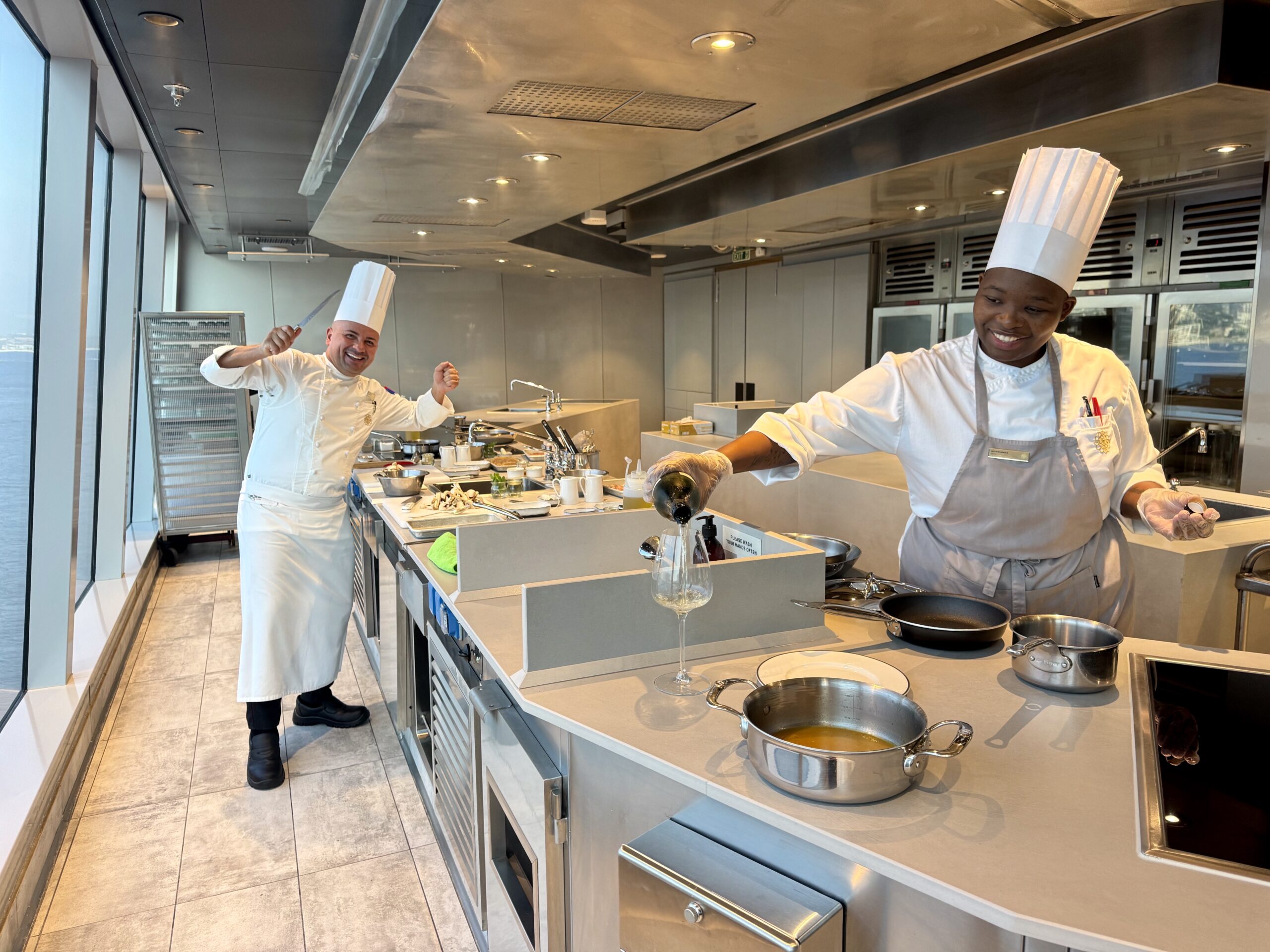 Two chefs in white uniforms and tall hats work in a stainless steel kitchen, one pouring wine into a pot while smiling at the camera.