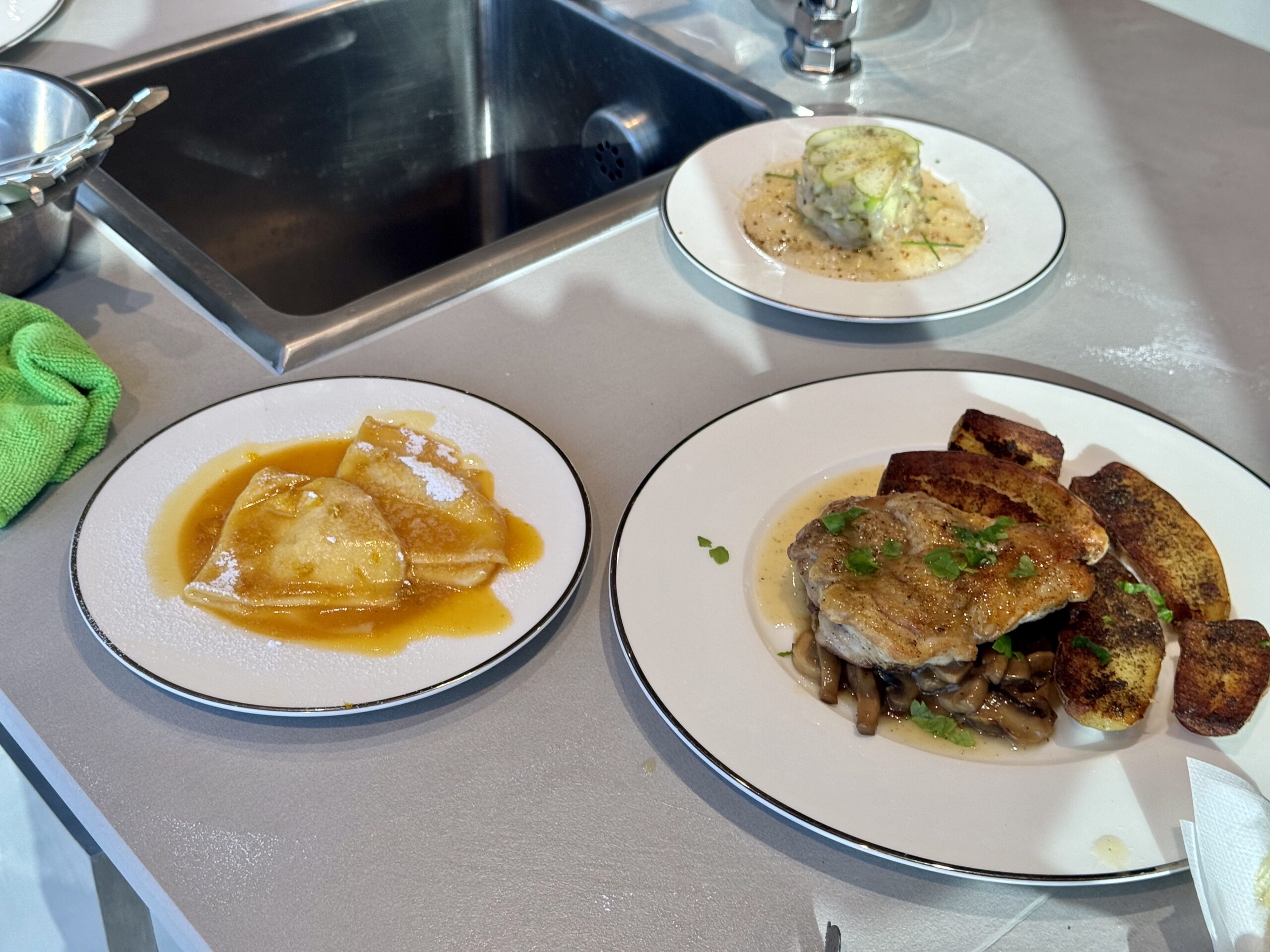 Three plated dishes on a stainless kitchen counter: a stacked cylinder of diced vegetables in a light sauce, with two other plates of food nearby (one with orange sauce and dumplings, another with meat and potatoes).