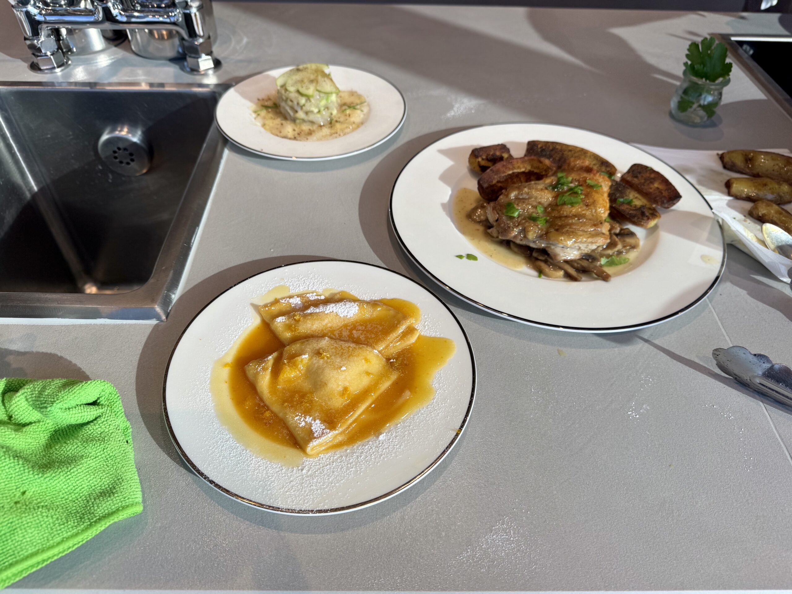 A bare kitchen counter with a sink on the left and plates of food: a white plate with stuffed ravioli in orange sauce in the foreground, and a larger plate with roasted chicken, potatoes, and gravy garnished with parsley on the right, plus a green cloth towel in the lower left corner and a small herb jar on the counter.