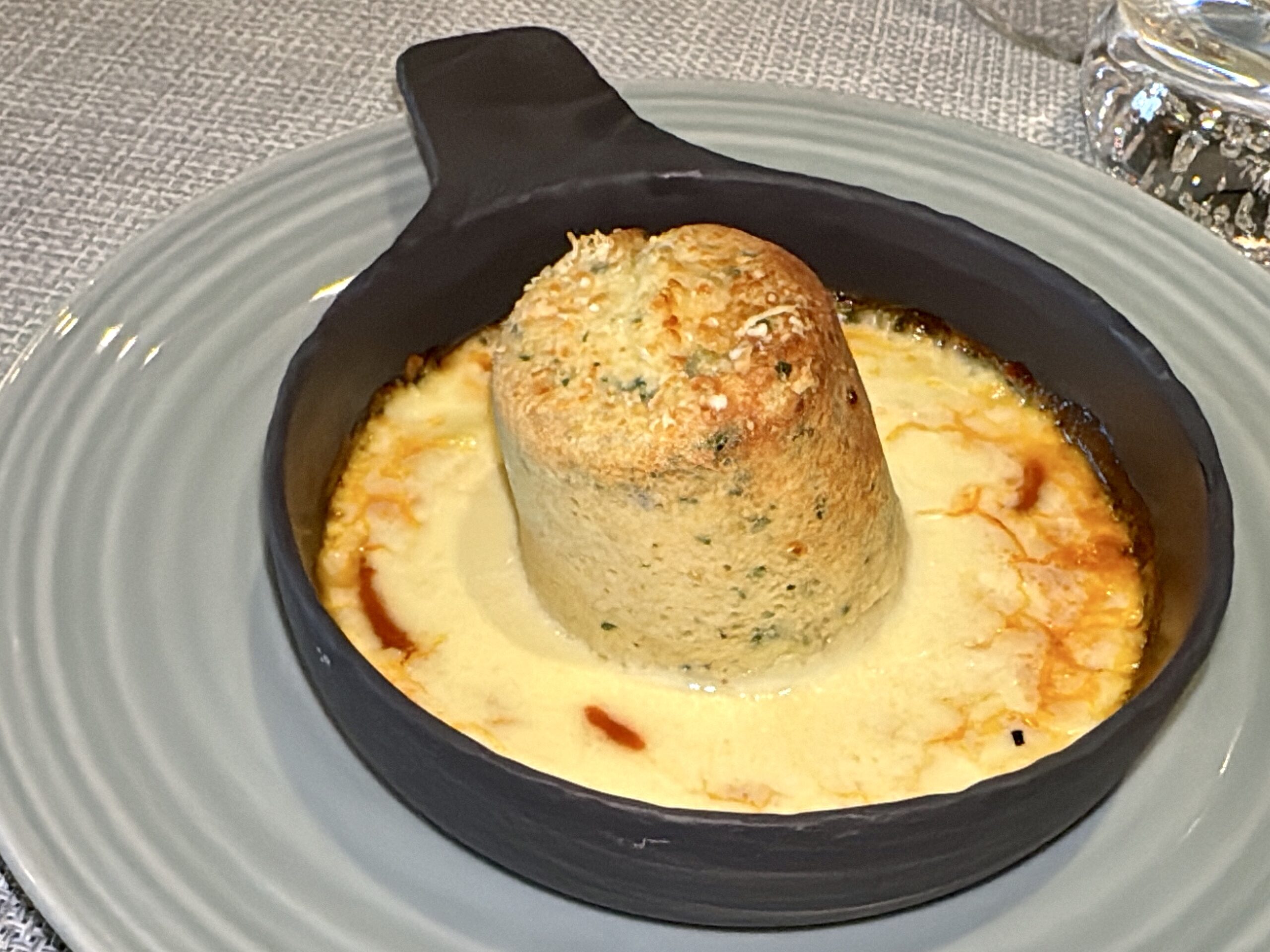 Small black cast-iron dish with a round herb loaf resting on creamy melted cheese sauce, on a gray plate.