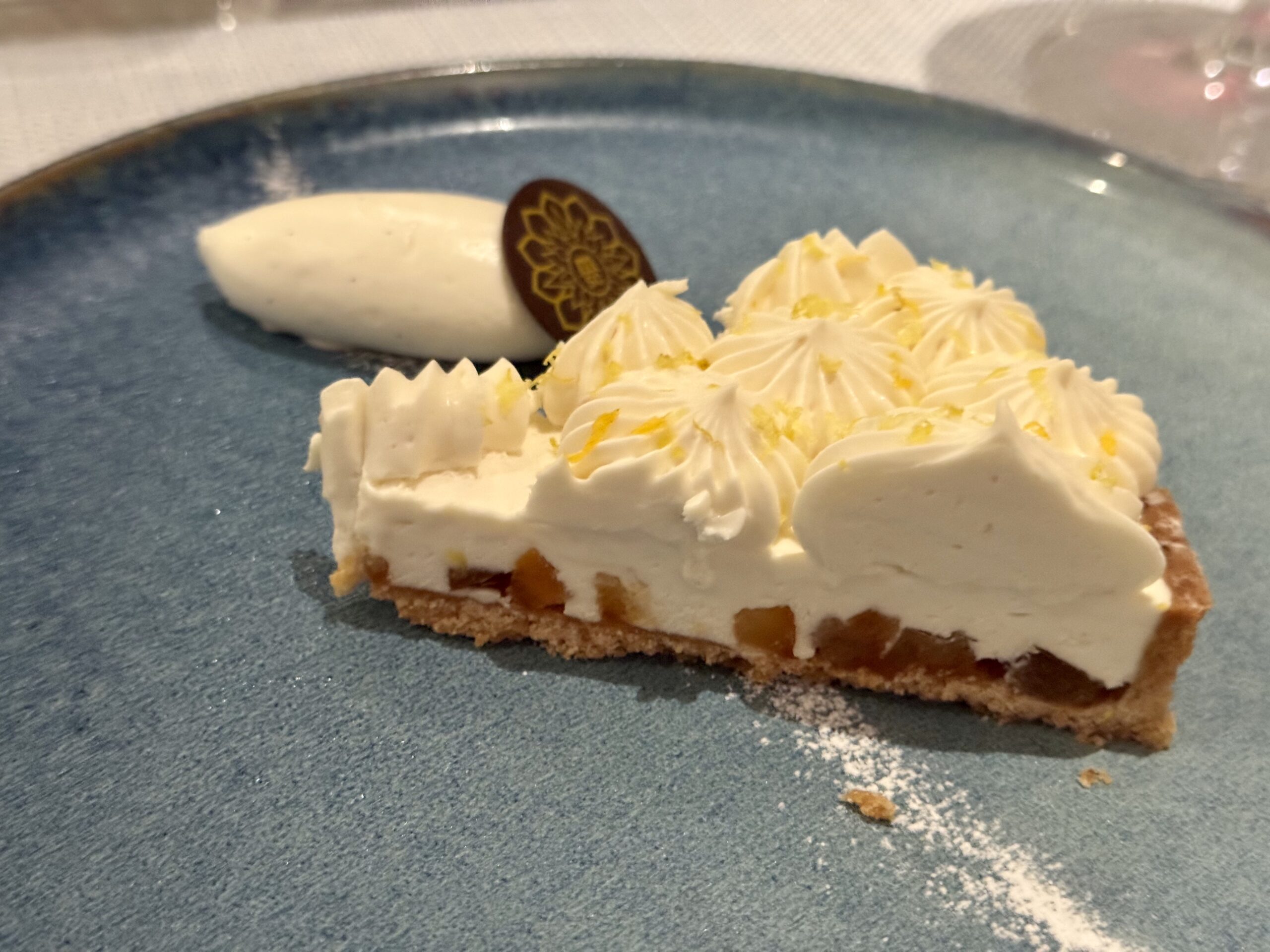 Slice of cheesecake on a blue plate with a decorative chocolate disk and swirls of whipped cream garnish on top.
