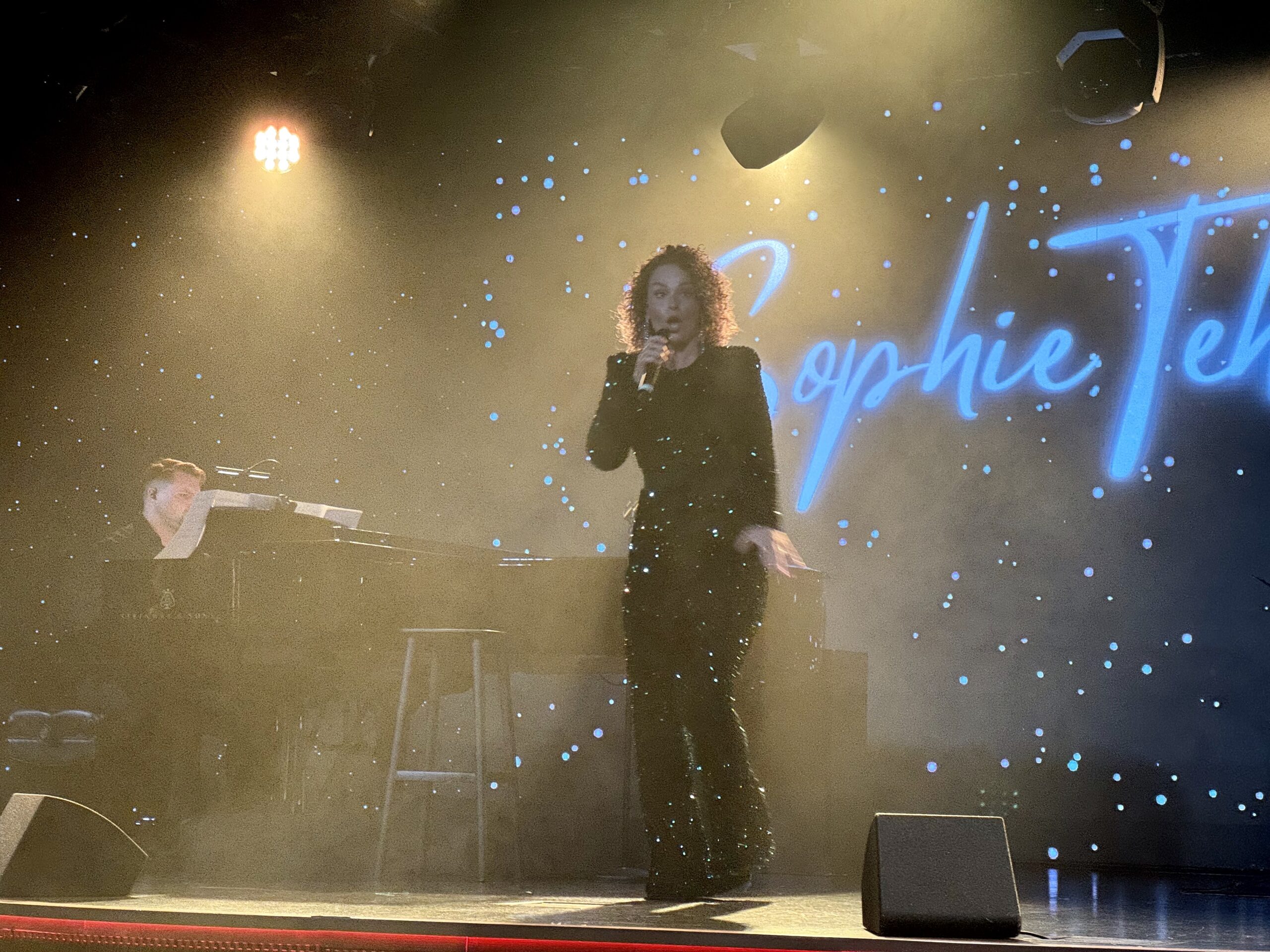 Singer with a handheld mic performing on stage, keyboardist visible to the left, and a starry blue-lit backdrop behind them.