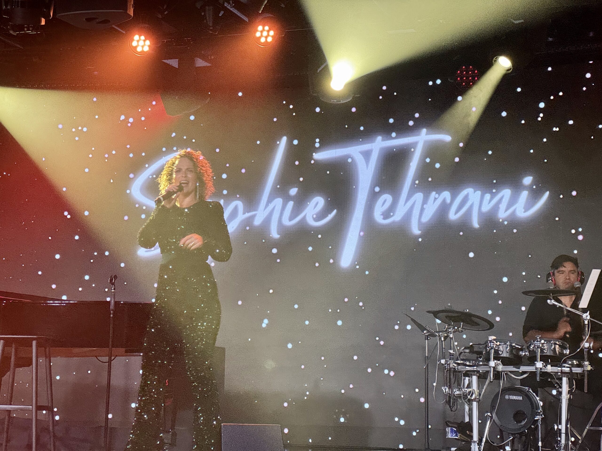 Female singer in a glittery outfit performs on stage with a drummer; orange/yellow spotlights and a starry backdrop.