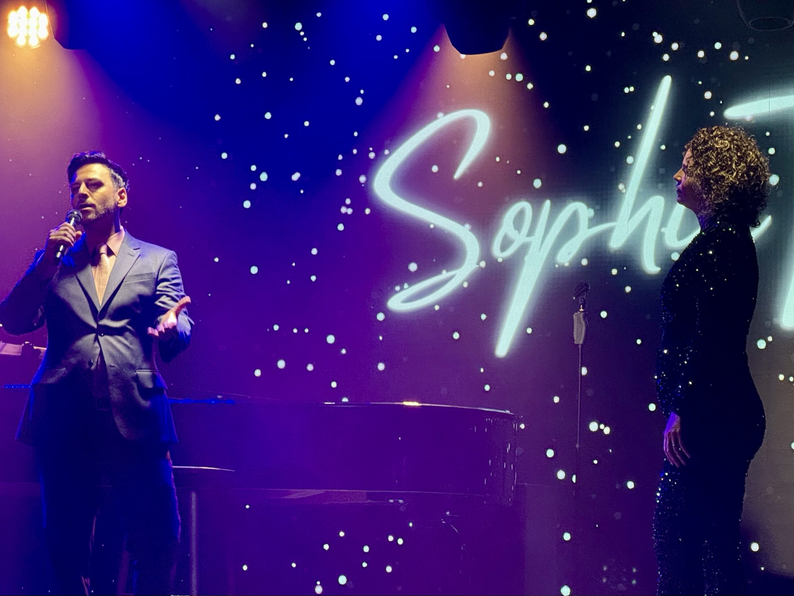 Male singer in a suit performing on a dimly lit stage with a neon script backdrop.