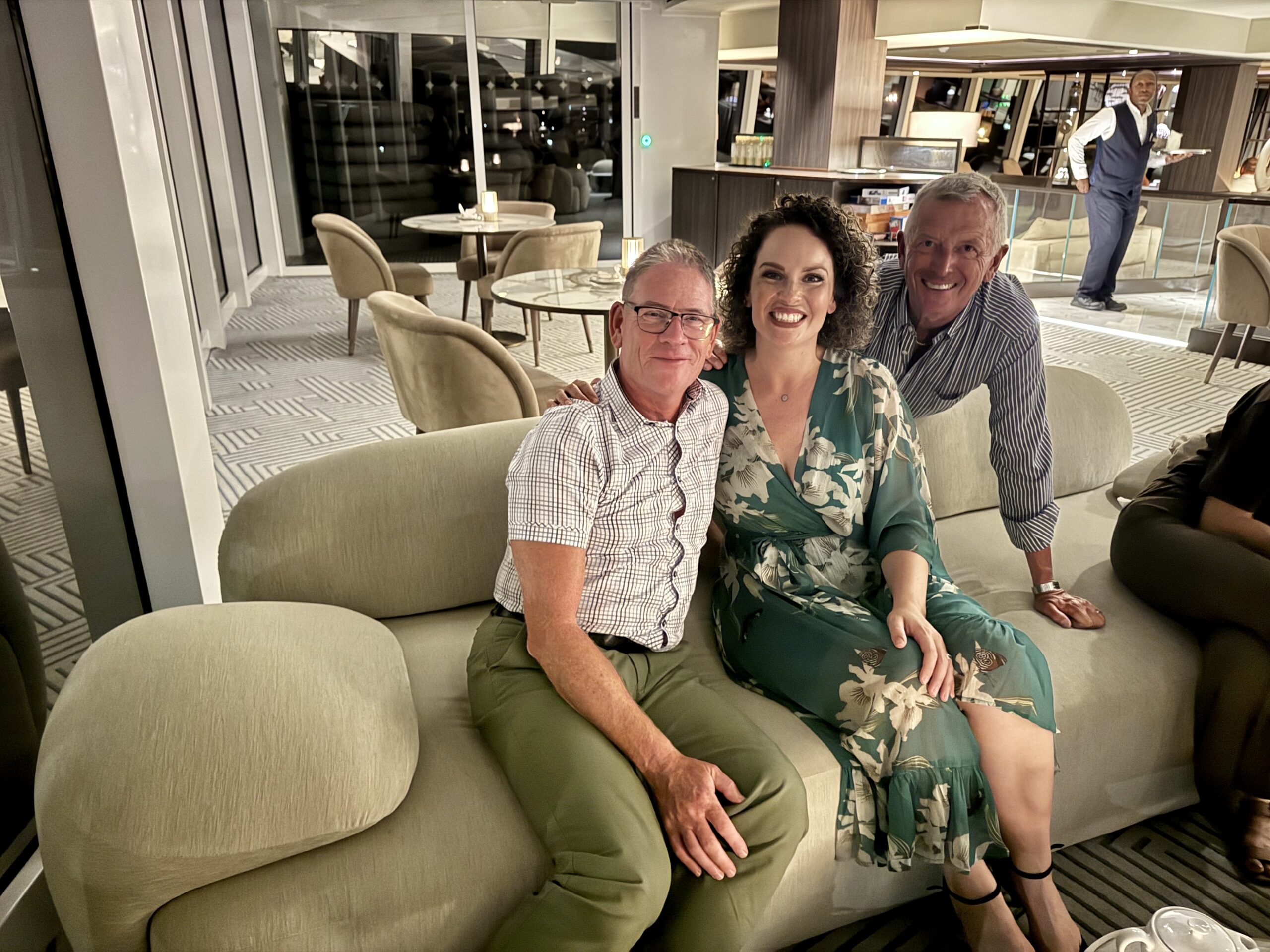 Three smiling adults sit close together on a cream sofa in a modern lounge; a server stands near the bar in the background.